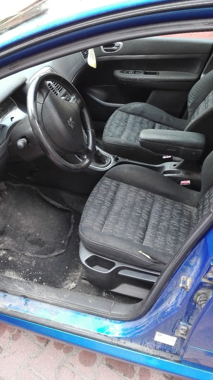 Wnętrze niebieskiego samochodu widziane z otwartych drzwi kierowcy. Widoczna jest czarna kierownica z logo marki Peugeot, dwa przednie fotele z ciemnoszarej tkaniny, podłokietnik oraz brudna podłoga z czarną wycieraczką.