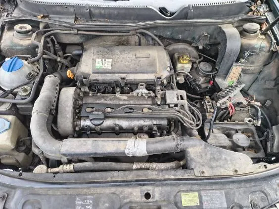 Widok na zanieczyszczony silnik samochodu z otwartą maską. W centralnej części znajduje się zasłonięty osłoną silnik z widocznymi przewodami, wężami i elementami mechanicznymi. Po lewej stronie widoczna niebieska pokrywka zbiornika, po prawej żółta pokrywka oraz różne przewody elektryczne. Na dole obrazu widoczne są części karoserii z niewyraźnymi etykietami. Całość pokryta jest kurzem i śladami zużycia.