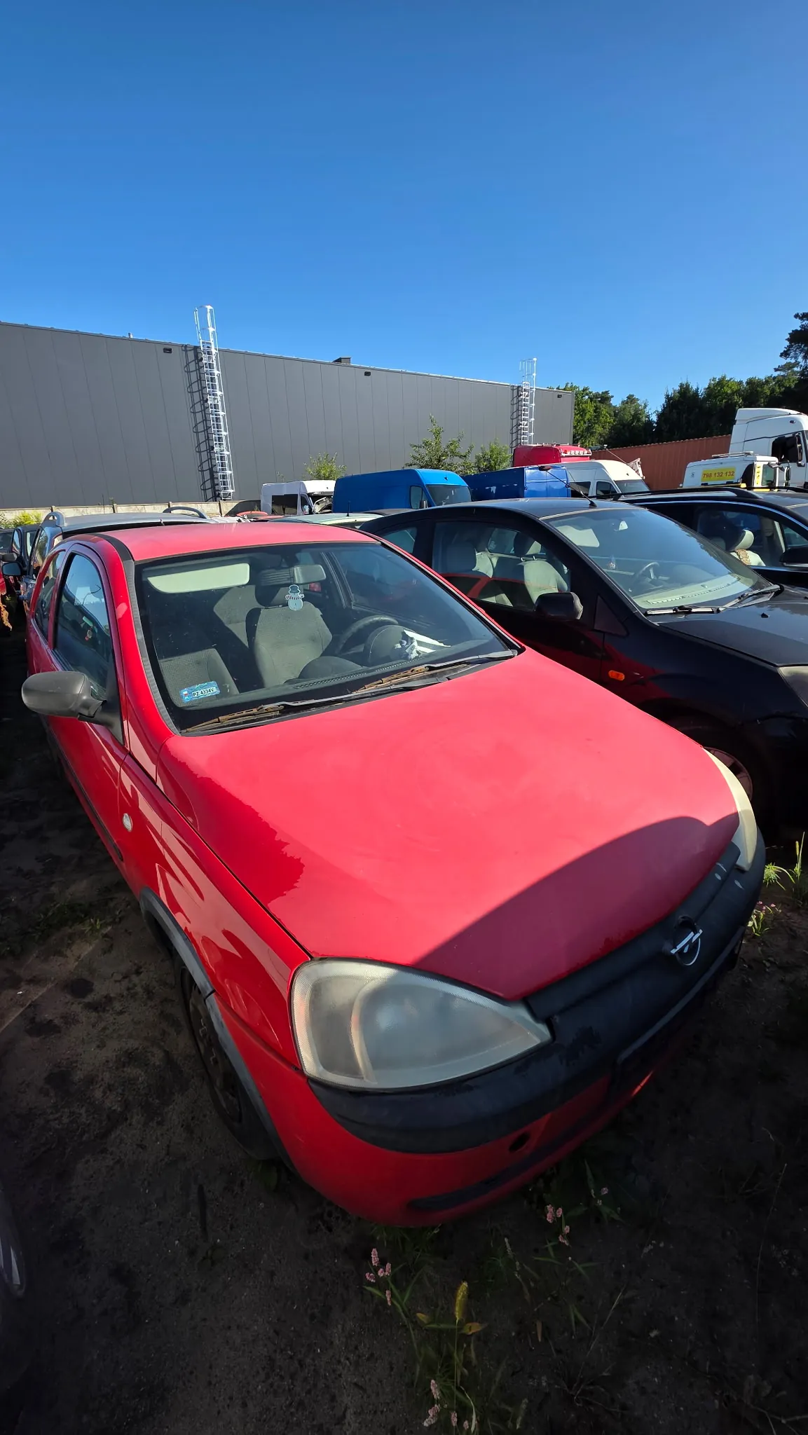 Czerwony samochód osobowy zaparkowany na placu obok innych pojazdów. W tle widoczny jest duży, szary budynek przemysłowy z zewnętrznymi schodami oraz niebieskie i białe furgonetki pod czystym, niebieskim niebem.