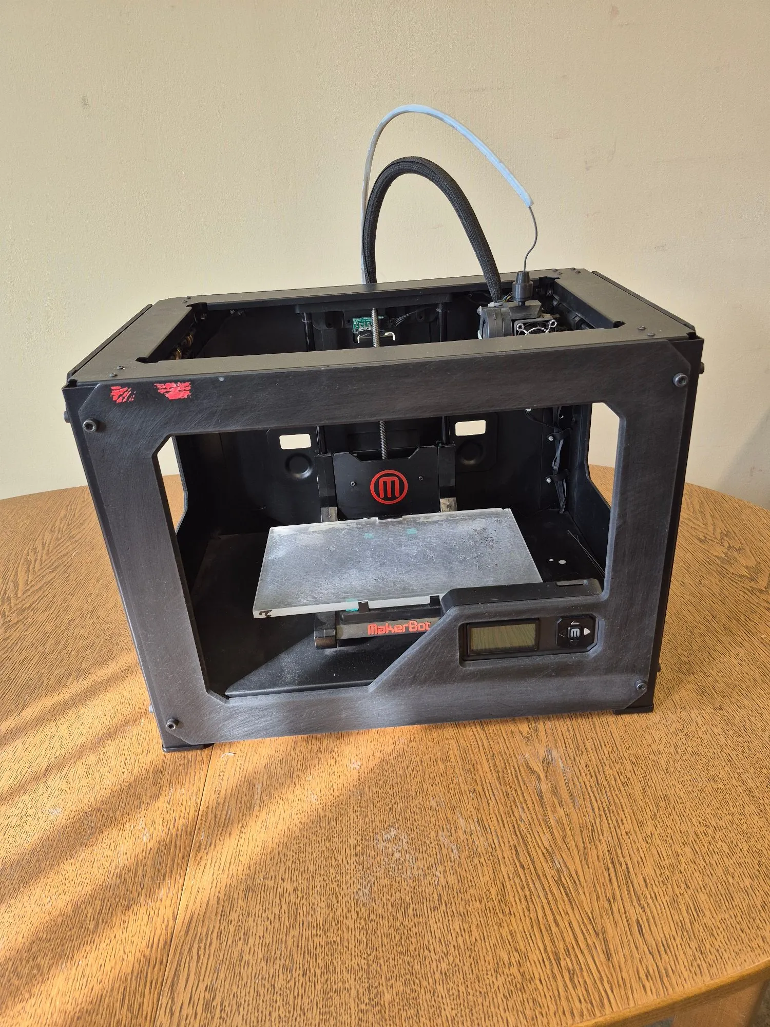 Czarna, otwarta obudowa drukarki 3D marki MakerBot, ustawiona na drewnianym stole. Wewnątrz widoczna jest platforma robocza oraz czerwone logo MakerBot w kształcie litery 'M' w okręgu. Na przednim panelu znajduje się napis 'MakerBot' oraz mały ekran LCD. Z górnej części urządzenia wystaje czarny przewód z niebieskim elementem zabezpieczającym.