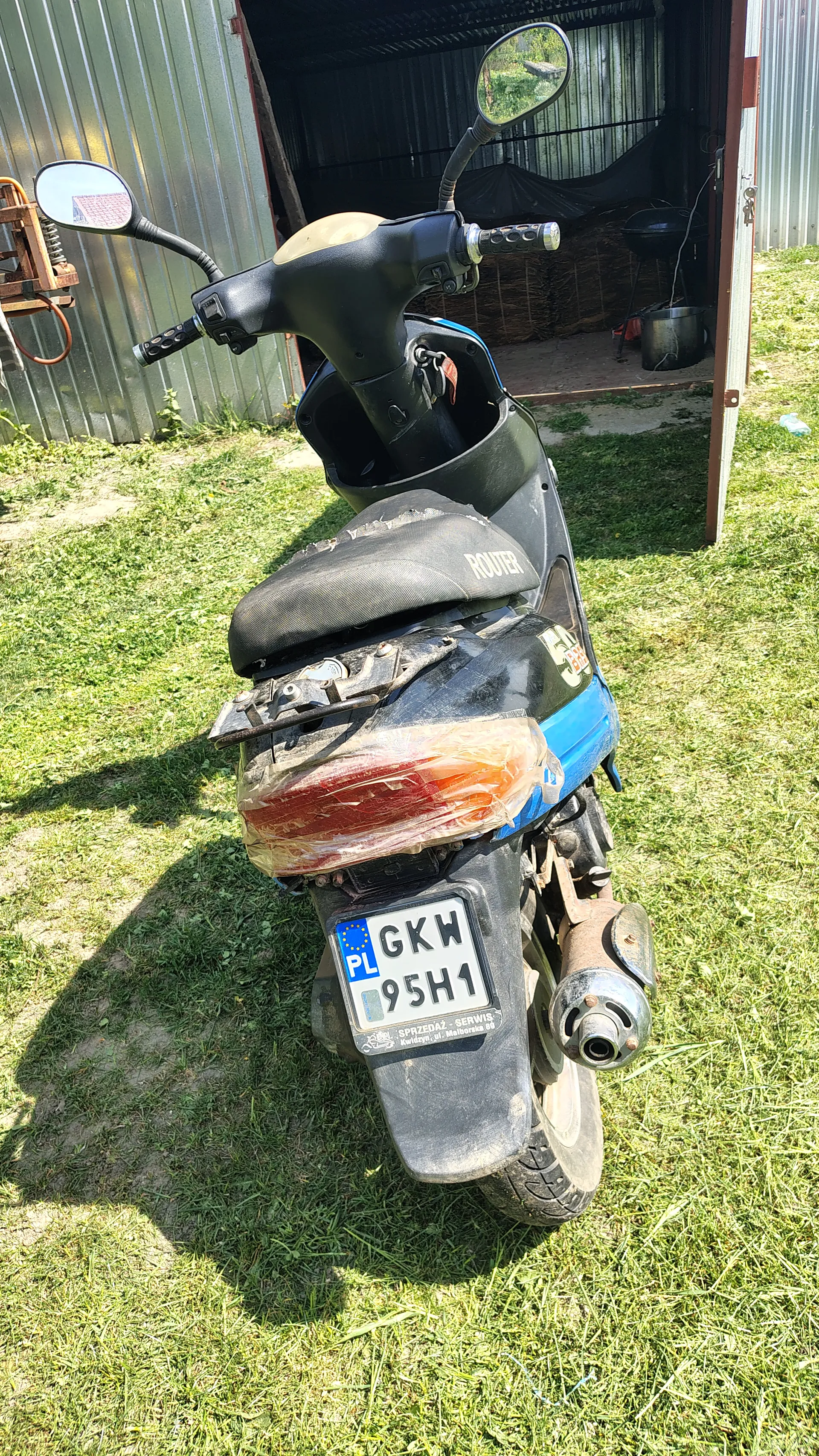 Czarno-niebieski skuter zaparkowany na trawie obok metalowego budynku. Widoczna tablica rejestracyjna z napisem 'PL GKM 95H1', tylna lampka owinięta przezroczystą folią, lusterka boczne oraz napis 'ROVER' na siedzeniu. W tle trawa i metalowa konstrukcja budynku.