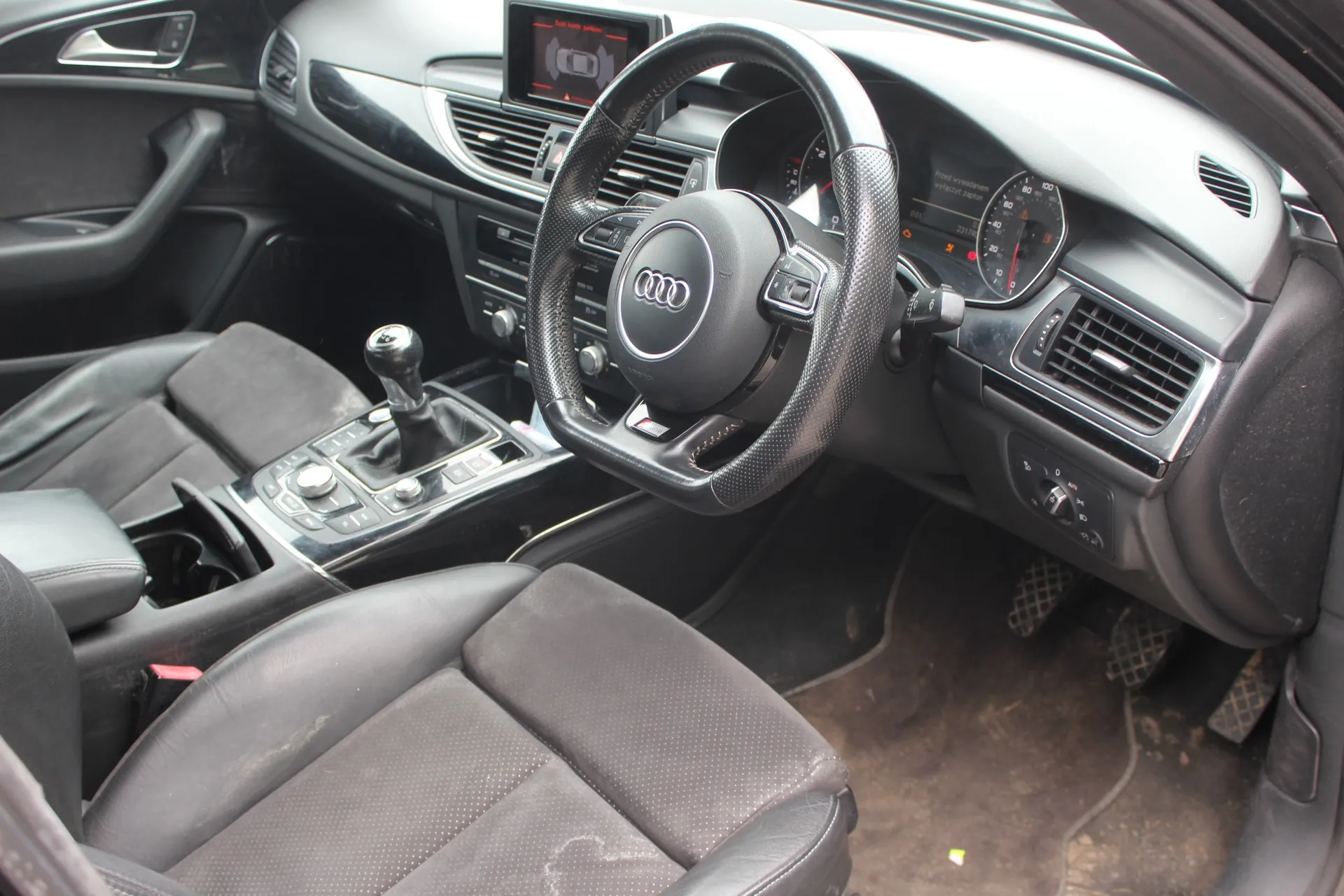 Wnętrze samochodu marki Audi, widok z perspektywy pasażera na stronę kierowcy. Widoczna jest skórzana kierownica z logo Audi, deska rozdzielcza z ekranem multimedialnym, zegary analogowe, lewarek zmiany biegów oraz dwa przednie fotele wykonane z czarnej skóry i materiału.
