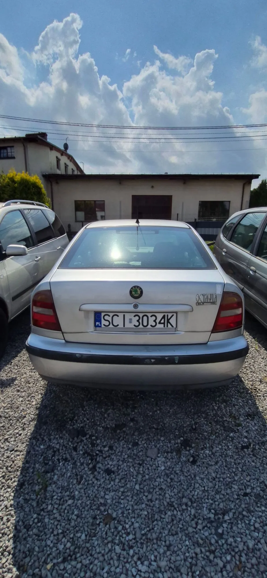 Srebrny samochód marki Skoda Octavia zaparkowany tyłem na żwirowym podjeździe. Na klapie bagażnika widoczna jest tablica rejestracyjna o numerze SCI 3034K oraz emblemat marki i napis OCTAVIA. Po lewej i prawej stronie znajdują się inne zaparkowane samochody. W tle widoczny jest niski, jasny budynek oraz fragment innego domu pod niebem z białymi chmurami.