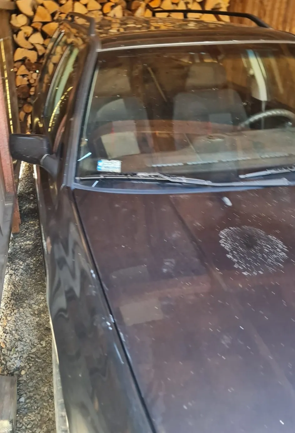 Zdjęcie przedstawia przód ciemnego samochodu zaparkowanego w pobliżu stosu drewna opałowego. Widoczna jest maska z widocznymi zabrudzeniami i odpryskami lakieru, przednia szyba z wycieraczkami oraz lusterko boczne. W tle widać ułożone polana drewna.