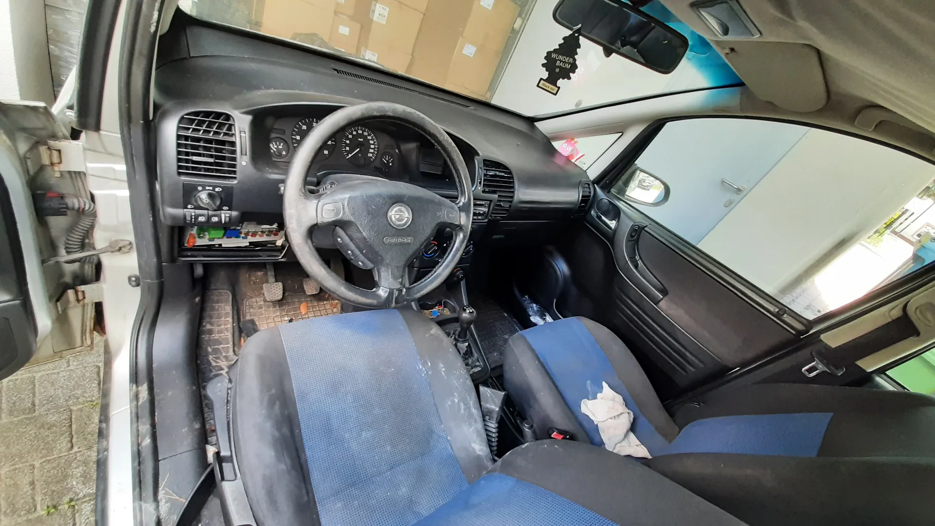 Wnętrze samochodu z widocznym kierownicą marki Opel, czarną deską rozdzielczą z licznikami, niebiesko-czarnymi siedzeniami w stanie zużytym. W otwartym drzwiach pasażera widoczna biała karoseria. Na lusterku wstecznym wiszący aromatyzator 'WUNDER-BAUM'. Podłoga pokryta brudem i liśćmi, widoczne przyciski klimatyzacji oraz fragmenty kabli pod deską rozdzielczą. Poza samochodem widoczne pudełka i biała ściana.