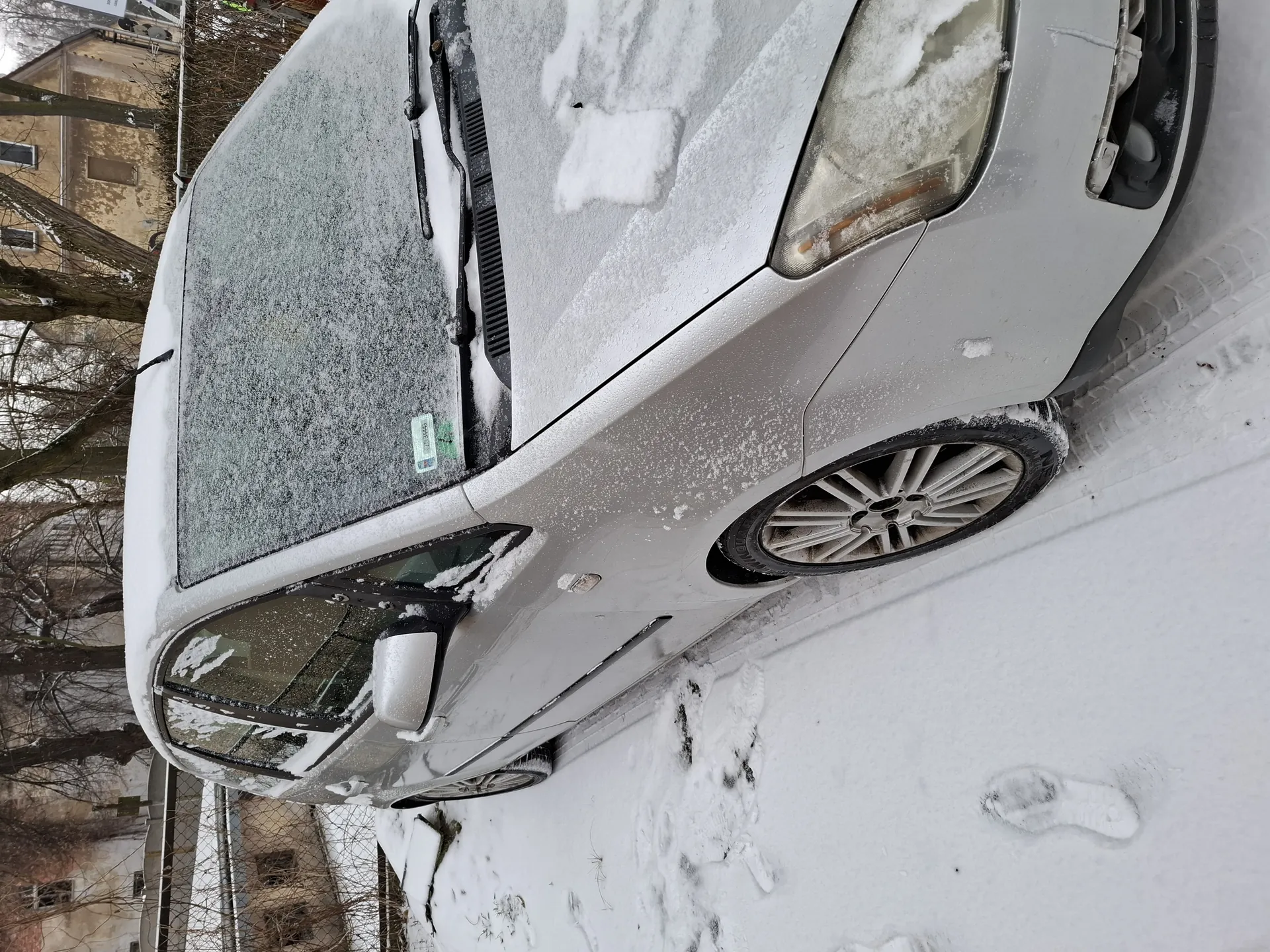 Srebrny samochód osobowy pokryty warstwą śniegu, zaparkowany na śnieżnej nawierzchni. Widoczne są odciski stóp w śniegu oraz ślady opon. Tło stanowią budynki, drzewa bez liści i ogrodzenie. Na przedniej szybie samochodu znajduje się nieczytelny naklejony dokument. Śnieg pokrywa dach, maskę i szyby pojazdu.