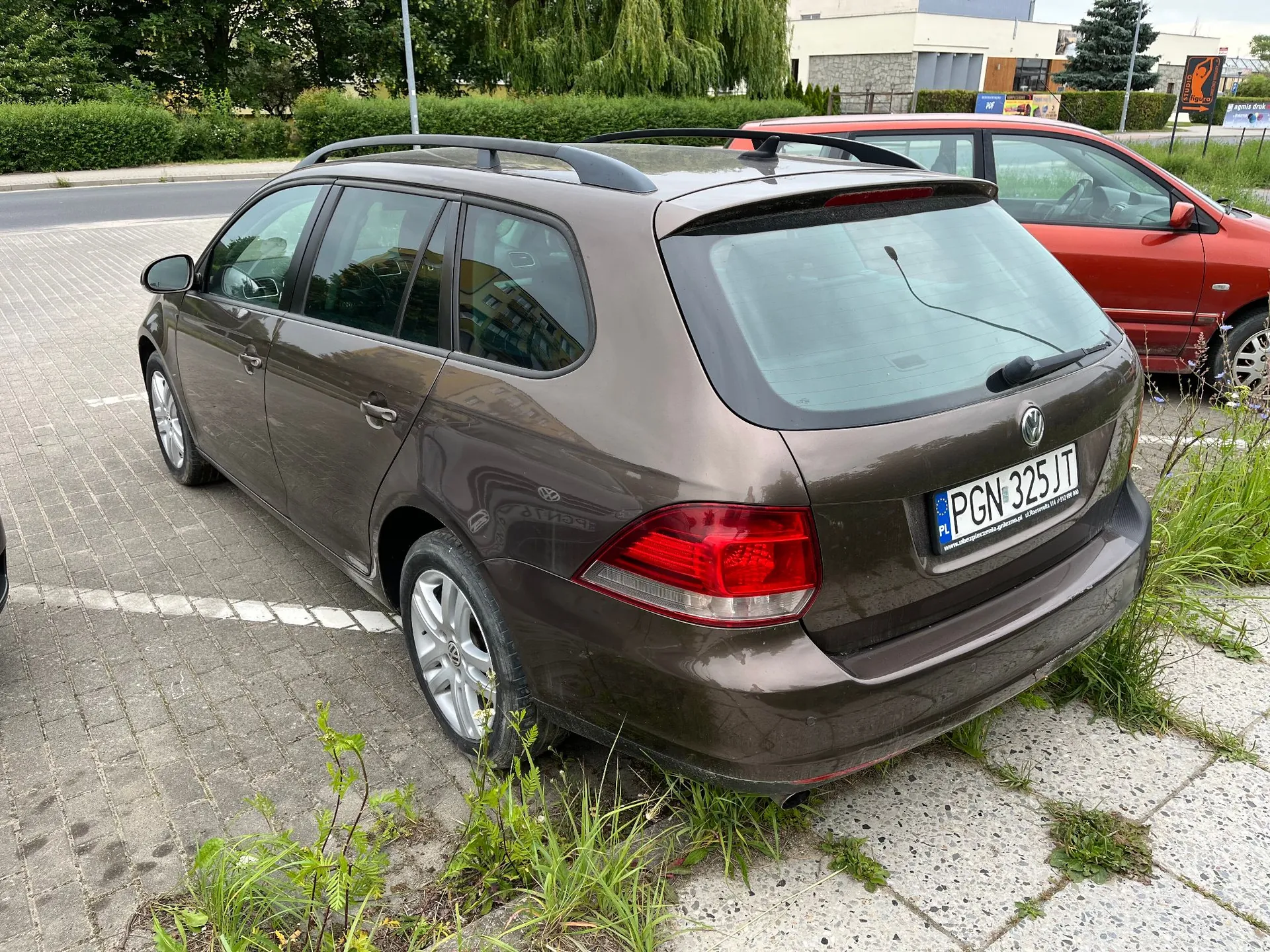 Brązowy samochód marki Volkswagen w nadwoziu kombi zaparkowany na kostce brukowej. Widoczna jest tylna część pojazdu z polską tablicą rejestracyjną o numerze PGN 325JT. W tle znajduje się czerwony samochód oraz zieleń i budynki.
