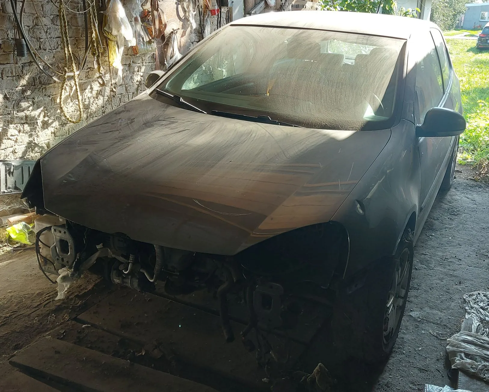 Zakurzony, ciemnoszary samochód osobowy typu hatchback zaparkowany w garażu lub warsztacie. Pojazd nie posiada przedniego zderzaka oraz reflektorów, odsłaniając elementy konstrukcyjne przodu. Karoseria jest pokryta grubą warstwą kurzu. W tle widoczna jest ceglana ściana z zawieszonymi narzędziami i szmatami.