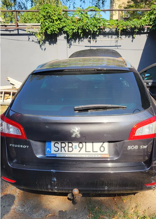 Tylny widok ciemnoszarego samochodu marki Peugeot 508 SW. Na klapie bagażnika znajduje się logo marki oraz tablica rejestracyjna o numerze SRB 9LL6 z oznaczeniem PL. Poniżej tablicy widnieje napis: PEUGEOT Ullarczyk Rybnik ul. Zielona 2 17 tel. 32/432 95 30 TOTAL. Samochód posiada hak holowniczy. W tle widać szary mur i zielone rośliny zwisające z ogrodzenia.