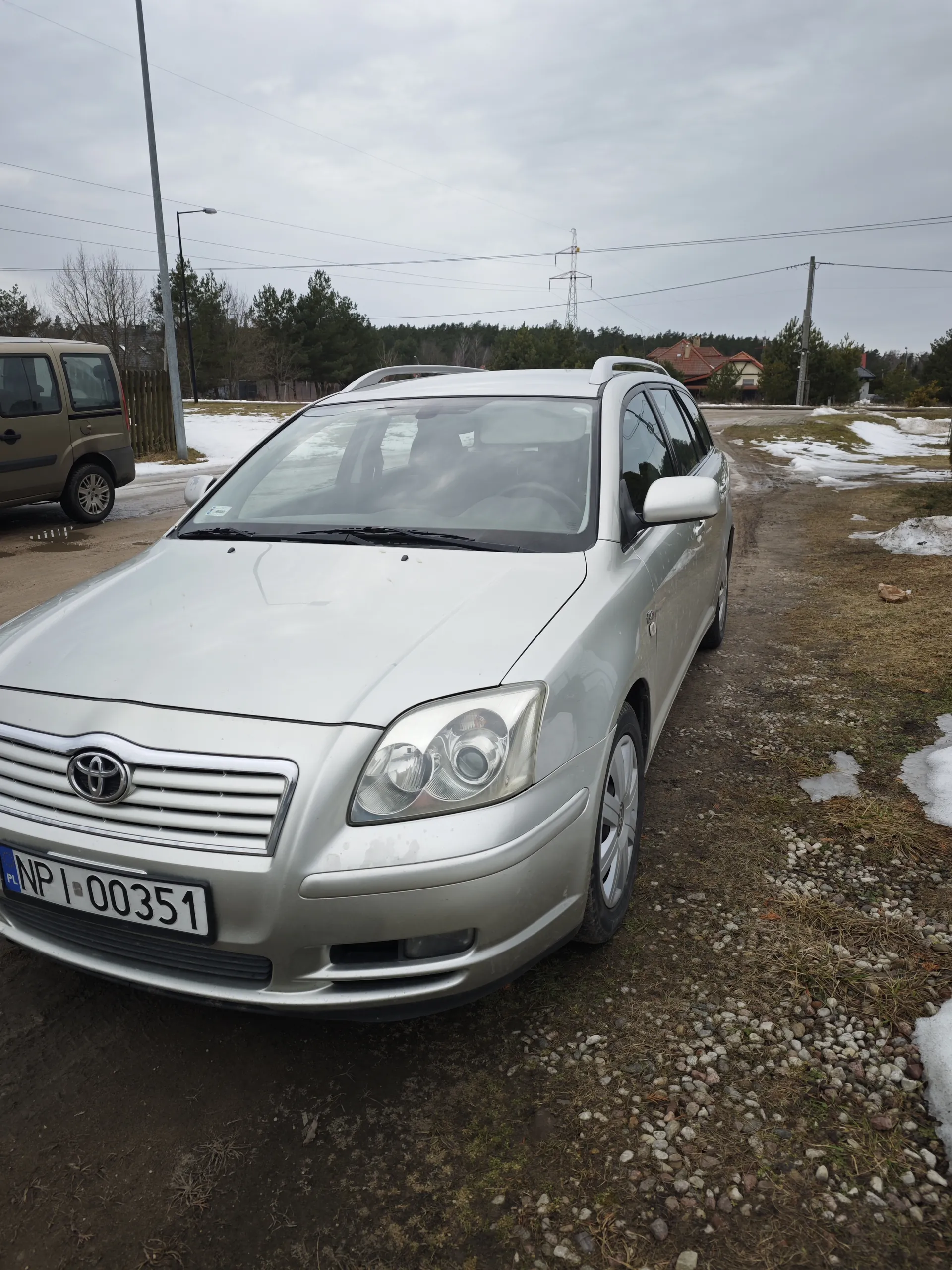 Srebrny samochód osobowy marki Toyota typu kombi z tablicą rejestracyjną PL NP 00351. Po lewej stronie brązowy van. Tło: droga żwirowa, resztki śniegu, drzewa, domy z czerwonymi dachami, słupy energetyczne z liniami, pochmurne niebo.