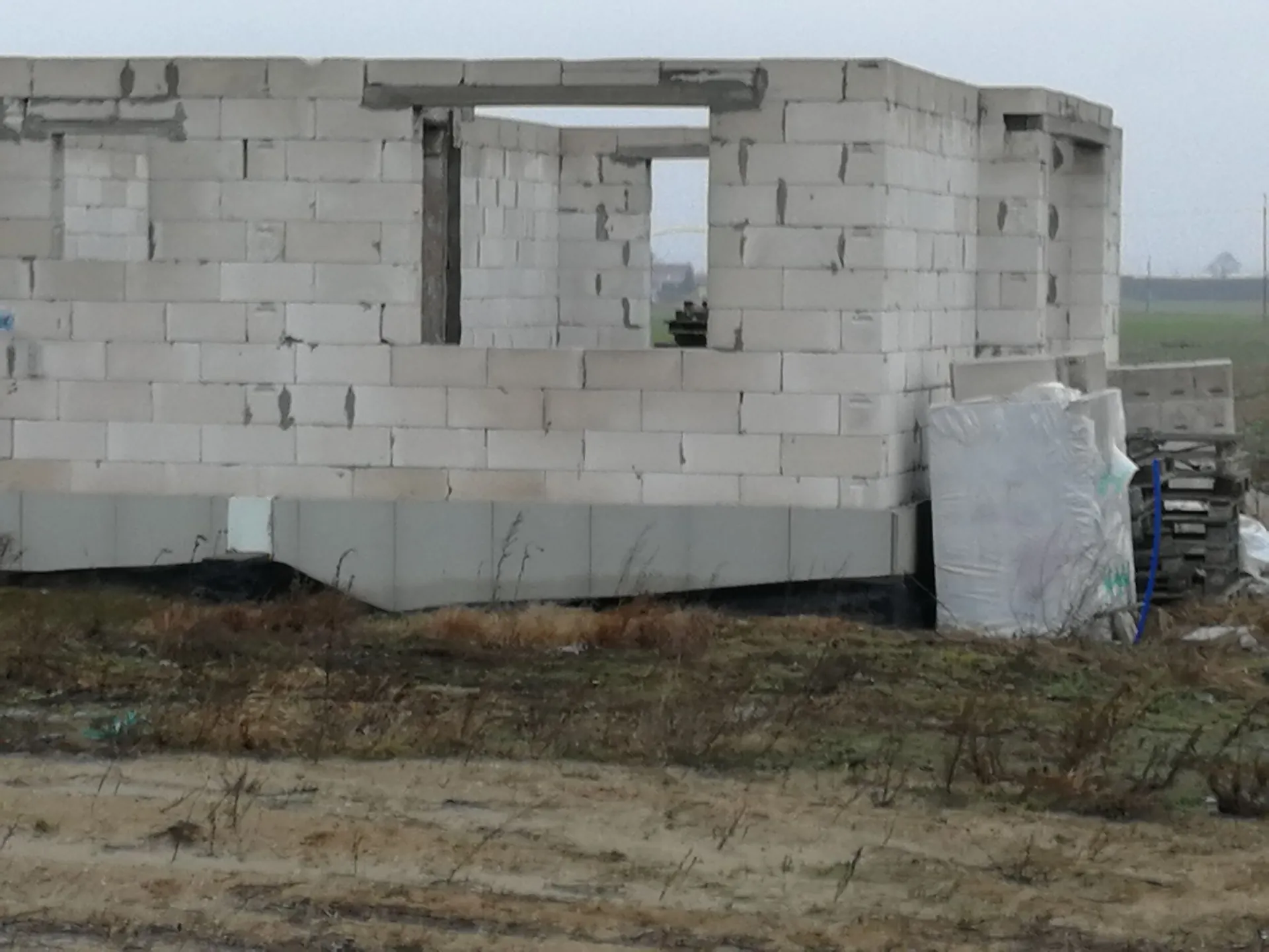 Zdjęcie przedstawiające budynek w stanie surowym, wzniesiony z jasnoszarych bloczków betonowych. Widoczne są otwory okienne oraz nadproża. Budynek stoi na betonowym fundamencie. Po prawej stronie znajduje się stos materiałów budowlanych przykrytych białą folią. W tle widać płaski, trawiasty teren pod szarym, zachmurzonym niebem.