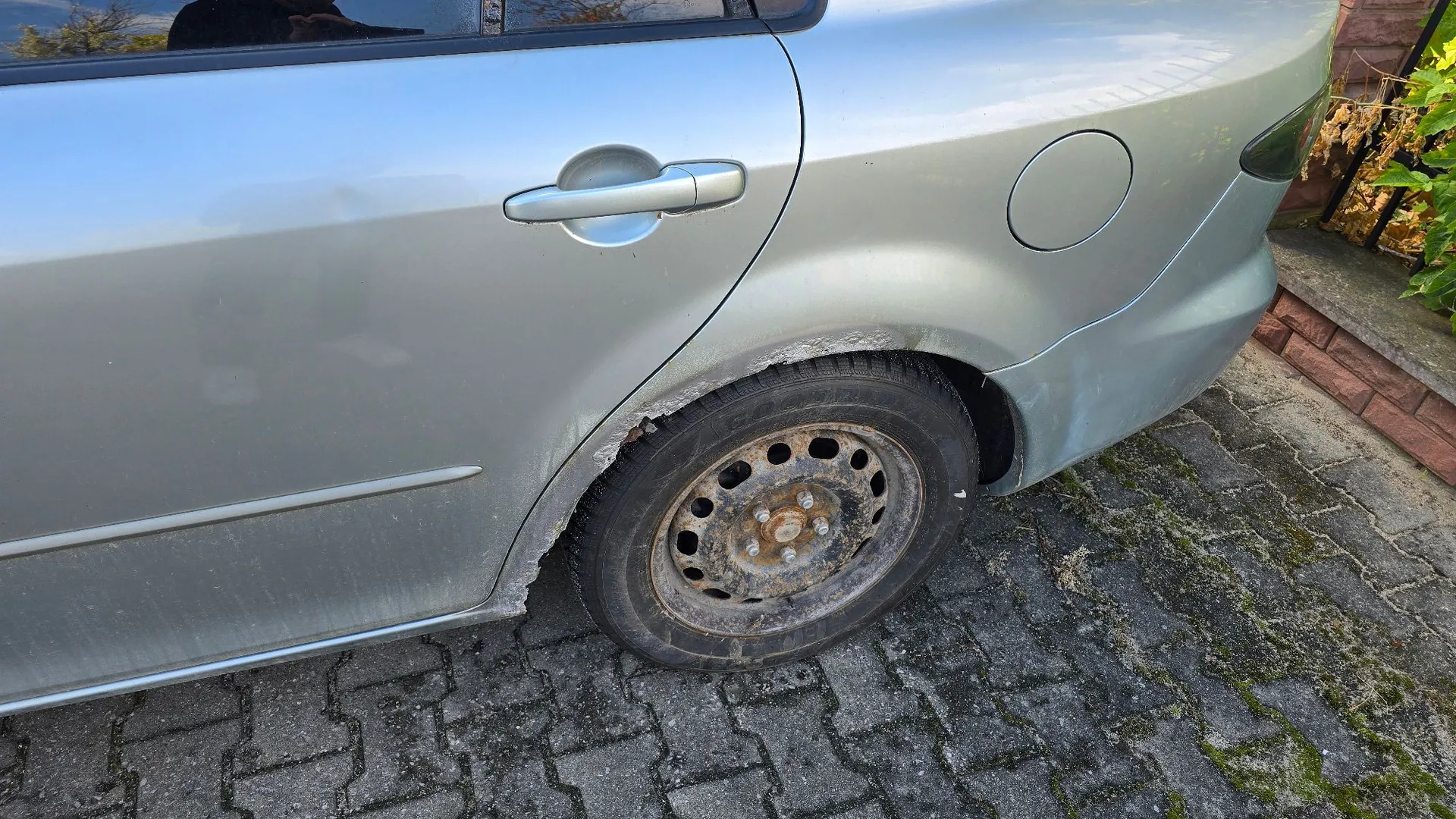 Zbliżenie na tylny prawy bok srebrnego samochodu. Widoczne jest uszkodzenie nadkola nad tylnym kołem w postaci rdzy i odprysków lakieru. Samochód ma stalową felgę z widocznymi śladami korozji i jest zaparkowany na kostce brukowej.