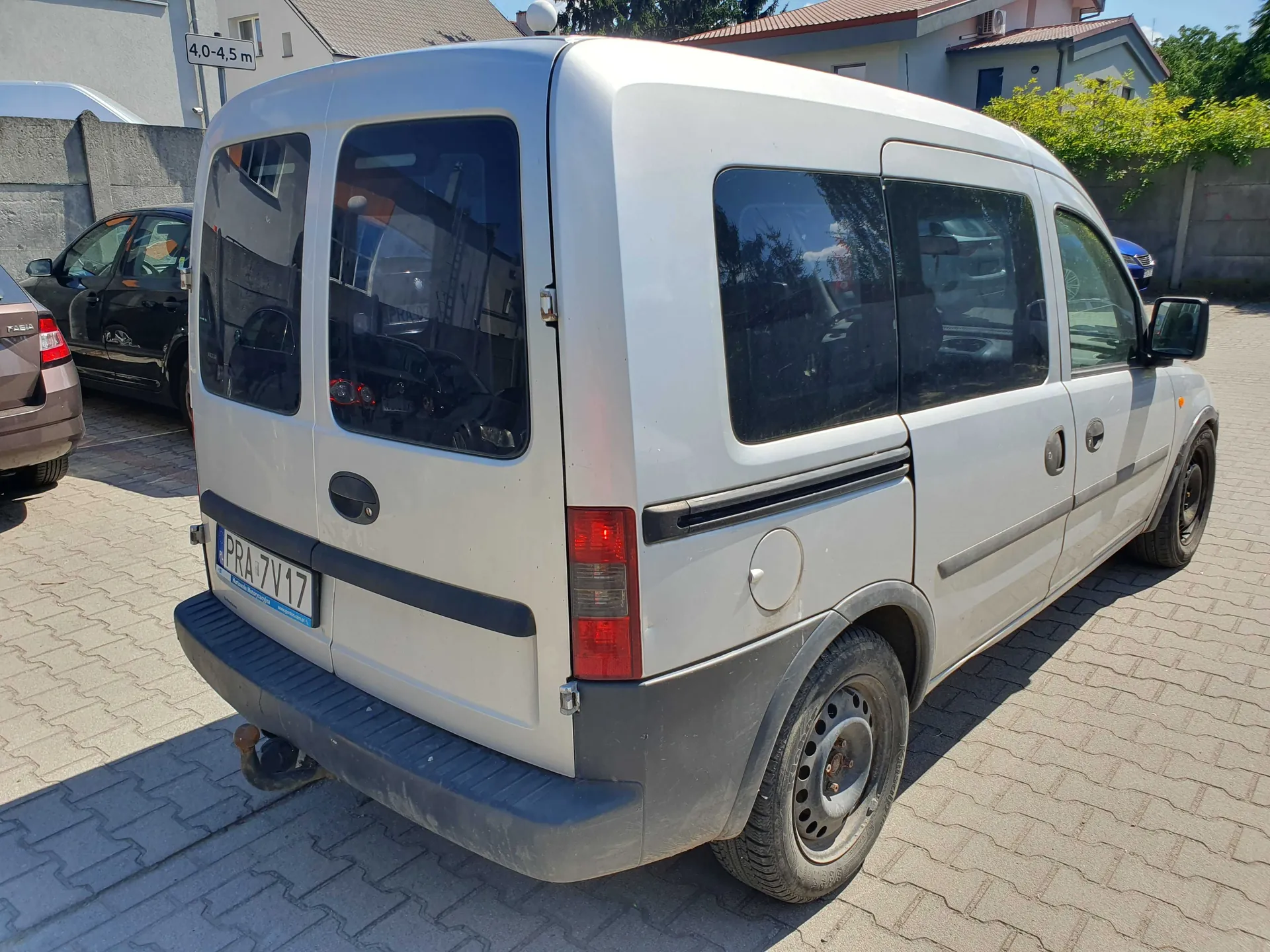 Srebrny samochód dostawczy typu van zaparkowany na kostce brukowej. Widok z tyłu i z boku pojazdu. Samochód ma czarny zderzak, czarne listwy boczne i stalowe felgi. Tablica rejestracyjna ma numer PRA 7V17. W tle widoczne są inne samochody, szary mur oraz fragmenty budynków mieszkalnych i zieleń.