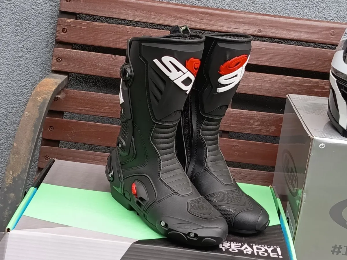 Para czarnych butów motocyklowych marki Sidi, ustawionych pionowo obok siebie na zielono-białym pudełku. Buty mają wysoki cholew z białym napisem 'SIDI' i czerwonym akcentem w górnej części. W tle widoczna jest drewniana ławka z brązowymi szczeblami oraz fragment białego kasku motocyklowego po prawej stronie.