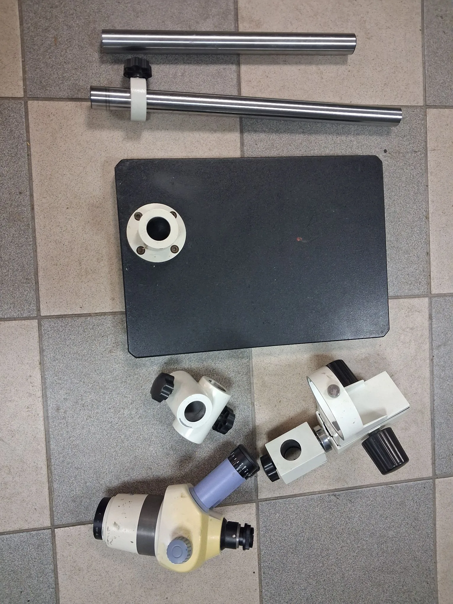 Zestaw metalowych i plastikowych elementów technicznych ułożonych na szarej, kafelkowej podłodze. W górnej części znajdują się dwa metalowe pręty, jeden z białym pierścieniem i czarną śrubą regulacyjną. Poniżej znajduje się czarna, prostokątna płyta z białym otworem montażowym. W dolnej części obrazu widoczne są białe i szare komponenty mechaniczne, w tym element przypominający uchwyt lub adapter z regulacyjnymi pokrętłami.