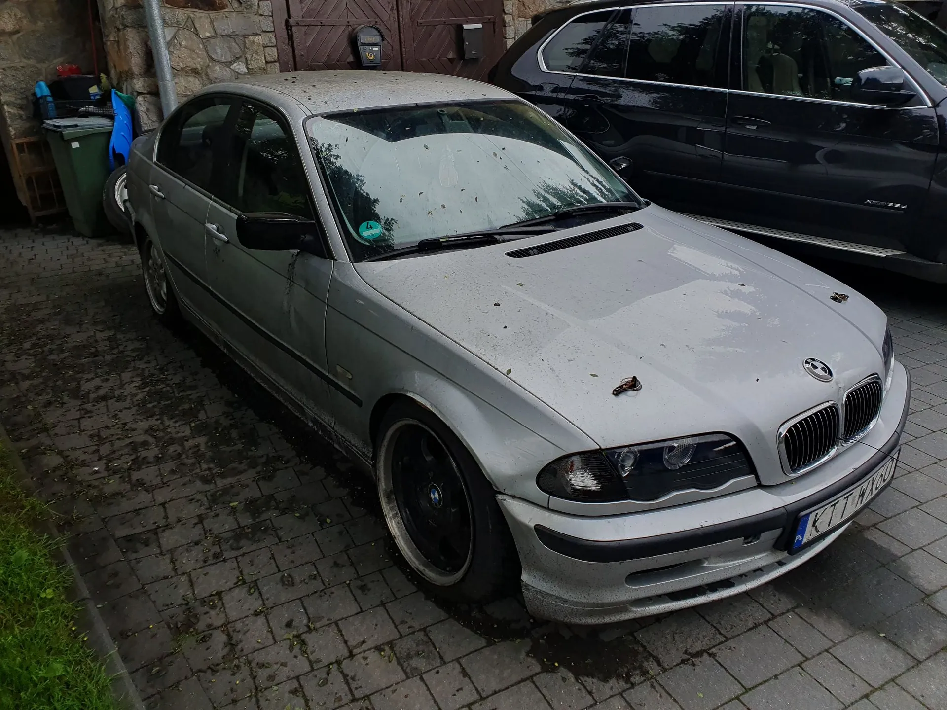 Srebrny samochód osobowy marki BMW serii 3 zaparkowany na kostce brukowej. Pojazd jest brudny, z widocznymi plamami i zaciekami na masce oraz przedniej szybie. W tle widoczny jest fragment ciemnego samochodu typu SUV, kamienna ściana budynku oraz zielony kosz na śmieci.