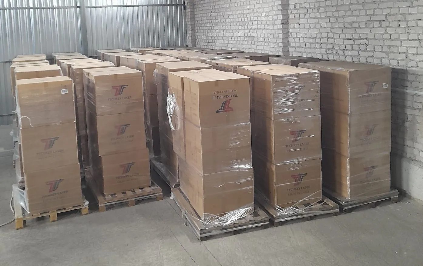 Wnętrze magazynu z ceglaną ścianą i metalowym poszyciem, w którym znajdują się liczne palety z brązowymi kartonami owiniętymi w folię stretch. Na kartonach widoczne jest czerwone logo oraz napis 'TECHKEY LASER MADE IN CHINA'.