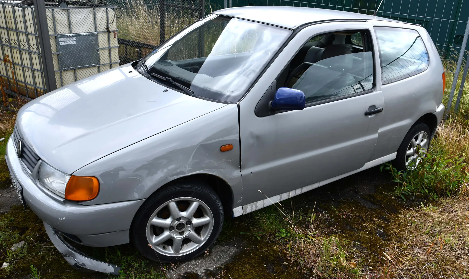 Srebrny samochód osobowy typu hatchback z uszkodzonym przodem i niebieskim lusterkiem bocznym. Pojazd stoi na nierównym terenie z trawą i mchem, w tle siatka ogrodzeniowa, przemysłowe zbiorniki oraz wysokie trawy. Widoczne są srebrne felgi, pomarańczowe kierunkowskazy i czarne siedzenia wewnątrz pojazdu.