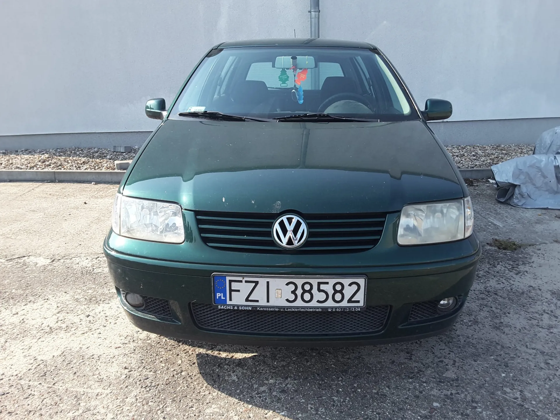 Widok z przodu ciemnozielonego samochodu marki Volkswagen zaparkowanego na żwirowym podłożu przed jasną ścianą budynku. Samochód ma polską tablicę rejestracyjną o numerze FZI 38582. Na masce widoczne jest logo Volkswagena, a wewnątrz auta, przy lusterku wstecznym, wisi kolorowa ozdoba.