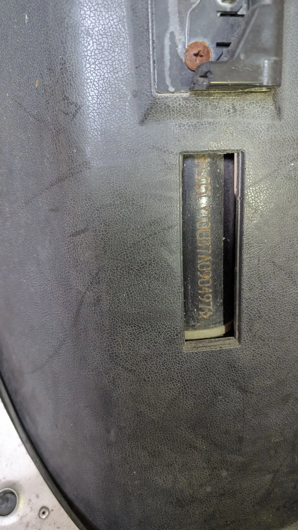 Zbliżenie na fragment czarnej, teksturowanej powierzchni z plastikowym wycięciem, w którym znajduje się pionowy pasek z nadrukowanym kodem seryjnym: 'S1580400377A0904974'. W górnej części zdjęcia widoczny jest fragment metalowego elementu przykręconego zardzewiałą śrubą.