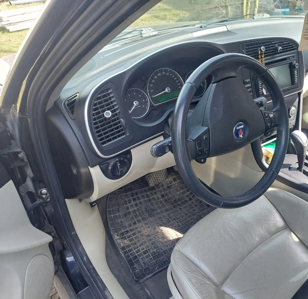 Wnętrze samochodu marki Ford widziane z perspektywy otwartych drzwi kierowcy. Widoczna jest czarna kierownica z logo Forda, deska rozdzielcza z zegarami, nawiewami powietrza oraz radiem. W stopie kierowcy znajduje się gumowa mata z wypustkami, a fotel jest wykonany z jasnej, beżowej skóry.