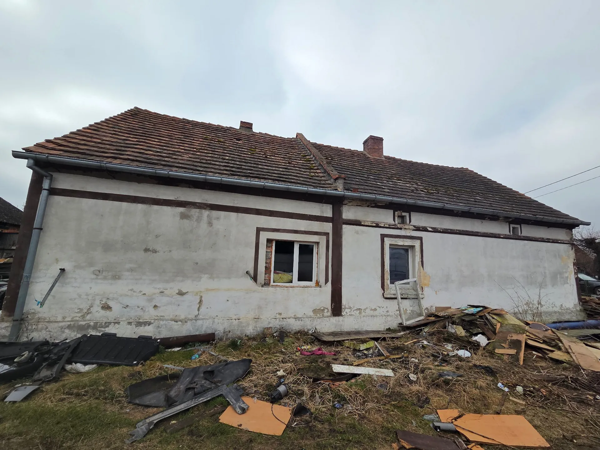 Zdjęcie starego, jednopiętrowego budynku z białymi, zniszczonymi tynkami i brązowym, dachówkowym dachem. Dom posiada dwa okna z brązowymi obramowaniami. Przed budynkiem znajduje się duże skupisko śmieci, w tym kawałki drewna, kartony, plastikowe elementy i gruz, rozrzucone na trawiastym terenie. Niebo jest zachmurzone i szare.