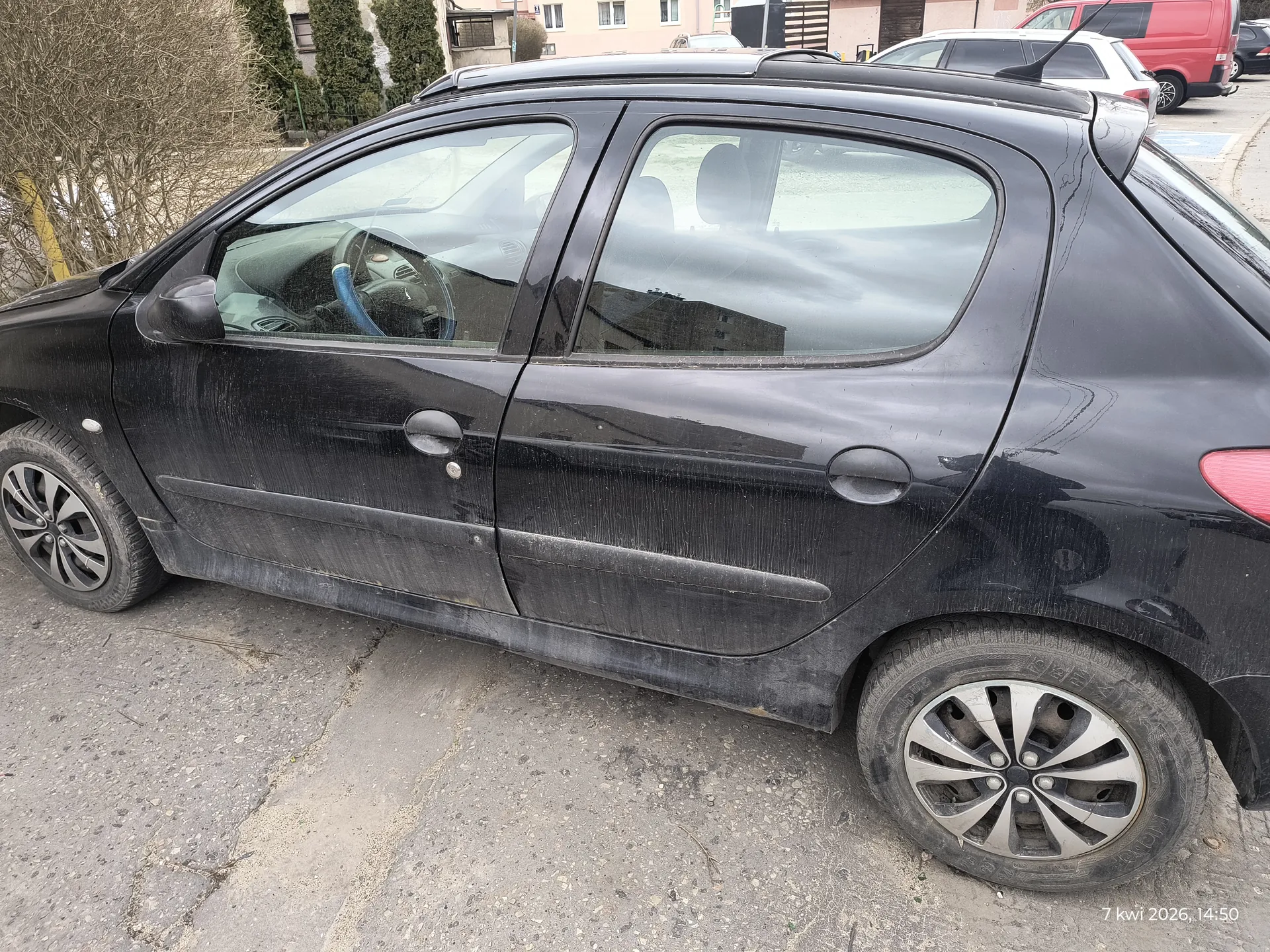 Czarny samochód osobowy typu hatchback zaparkowany na asfaltowej nawierzchni. Zewnętrzne elementy pojazdu są zabrudzone, widoczne ślady kurzu i brudu na karoserii oraz kołach. Wewnątrz samochodu kierownica z niebieskim akcentem. W tle inne pojazdy: biały samochód i czerwony van, budynki mieszkalne z oknami oraz krzewy. Na dole obrazu widoczny napis: 7 kwi 2026, 14:50