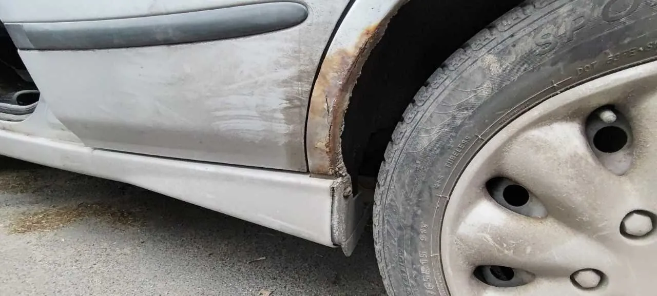 Zbliżenie na przednią prawą część nadwozia samochodu w kolorze srebrnym. Widoczna jest zardzewiała obudowa koła, brudna opona z napisem 'SP' oraz felga z pięcioma otworami. Nadwozie pokryte jest zabrudzeniami, a w tle asfaltowa powierzchnia z drobnymi śmieciem.