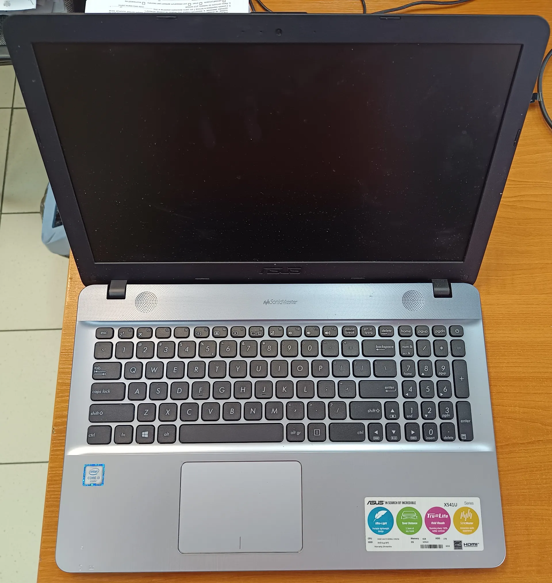 Otwarty laptop marki ASUS model X541U z czarnym ekranem w stanie wyłączonym. Widoczna klawiatura z czarnymi klawiszami, szary touchpad oraz naklejki: Intel Core i3 w lewym dolnym rogu, a w prawym dolnym rogu zestaw naklejek ASUS z napisami 'In Search of Incredible', 'X541U', 'TruLife' oraz ikonami technologii. Laptop umieszczony na drewnianym biurku, w tle fragment podłogi z białymi płytkami i częściowo widoczne przewody. Na górze obrazu widoczny fragment białego dokumentu.