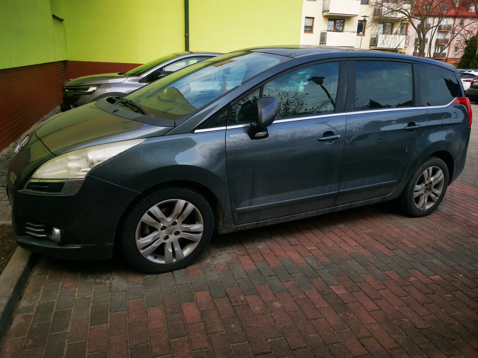 Ciemnoszary samochód typu minivan zaparkowany na ceglanym chodniku. Po lewej stronie widoczna zielona i czerwona ściana budynku, w tle częściowo widoczny srebrny samochód oraz białe i różowe budynki mieszkalne z oknami i balkonami. Na przednim szybie odbijają się drzewa bez liści.