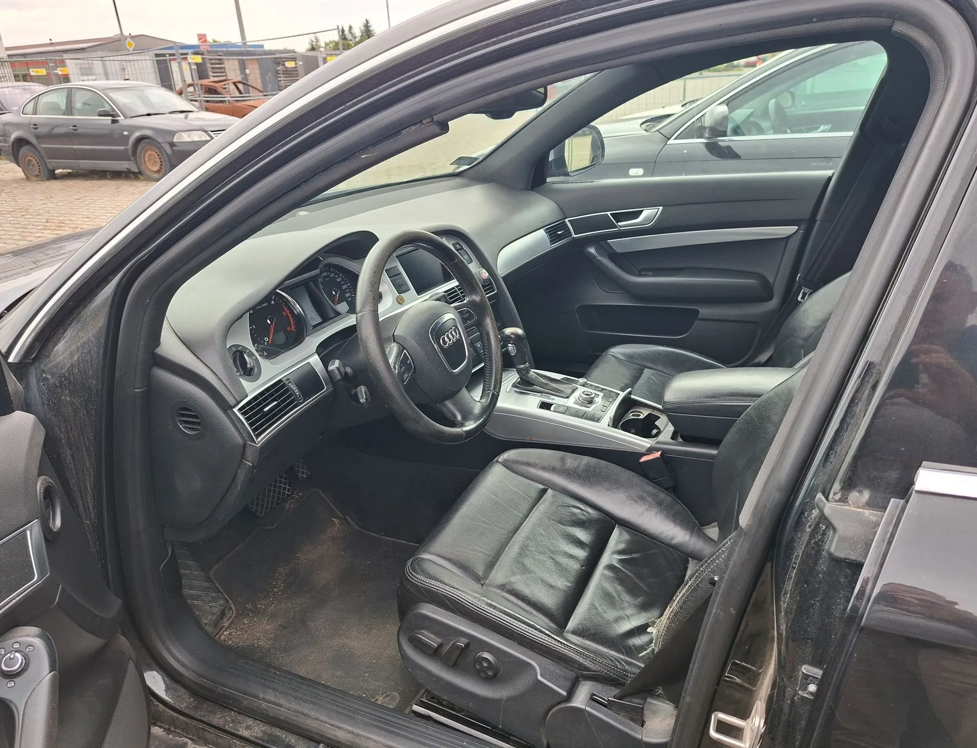 Widok z otwartych drzwi pasażera na wnętrze czarnego samochodu marki Audi. Widoczna jest czarna skórzana tapicerka foteli, kierownica z logo Audi, deska rozdzielcza z zegarami oraz konsola środkowa. W tle, za szybą, widoczny jest inny szary samochód zaparkowany na zewnątrz.