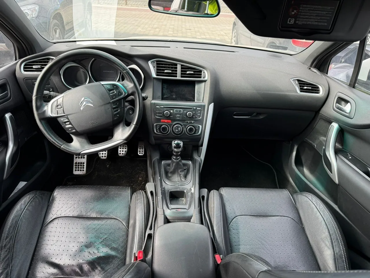 Wnętrze samochodu Citroën z widokiem na deskę rozdzielczą, kierownicę z logo marki, centralny panel z ekranem i przyciskami, dźwignię zmiany biegów oraz czarne skórzane fotele z widocznymi śladami zużycia. Przez przednią szybę widoczny parking z innymi pojazdami. W tylnej części deski rozdzielczej znajduje się lusterko wsteczne oraz tabliczka z informacjami.