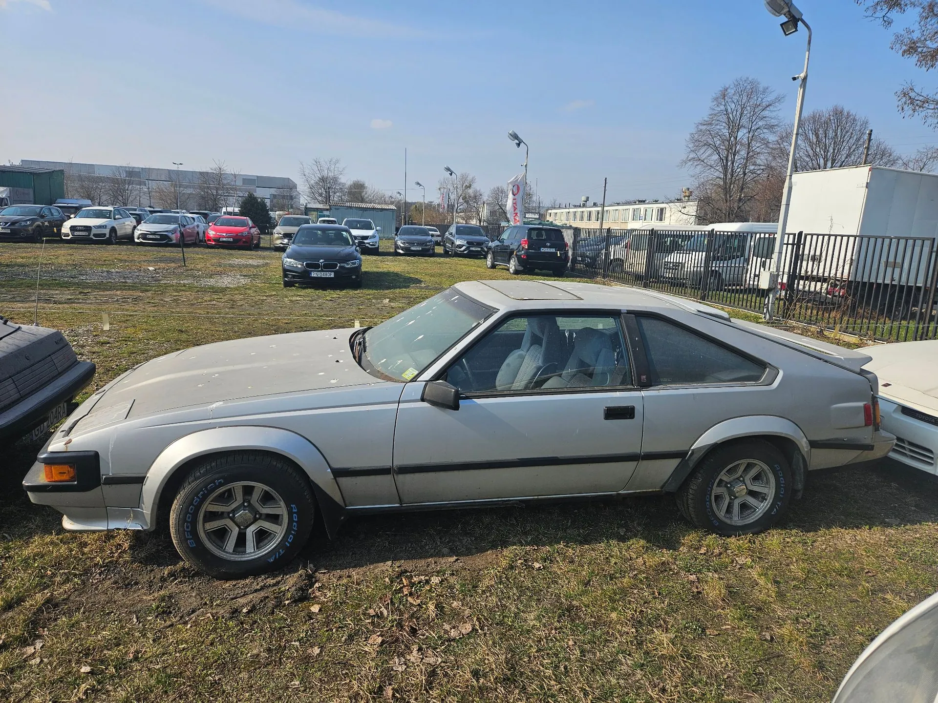 Srebrny, dwudrzwiowy samochód osobowy typu coupe z lat 80. zaparkowany na trawiastym placu. Pojazd jest widoczny z profilu, ma charakterystyczną kanciastą sylwetkę i ciemne felgi z białymi napisami na oponach. W tle znajduje się parking z wieloma innymi samochodami, ogrodzenie z czarnych prętów oraz białe budynki przemysłowe pod jasnym, niebieskim niebem.