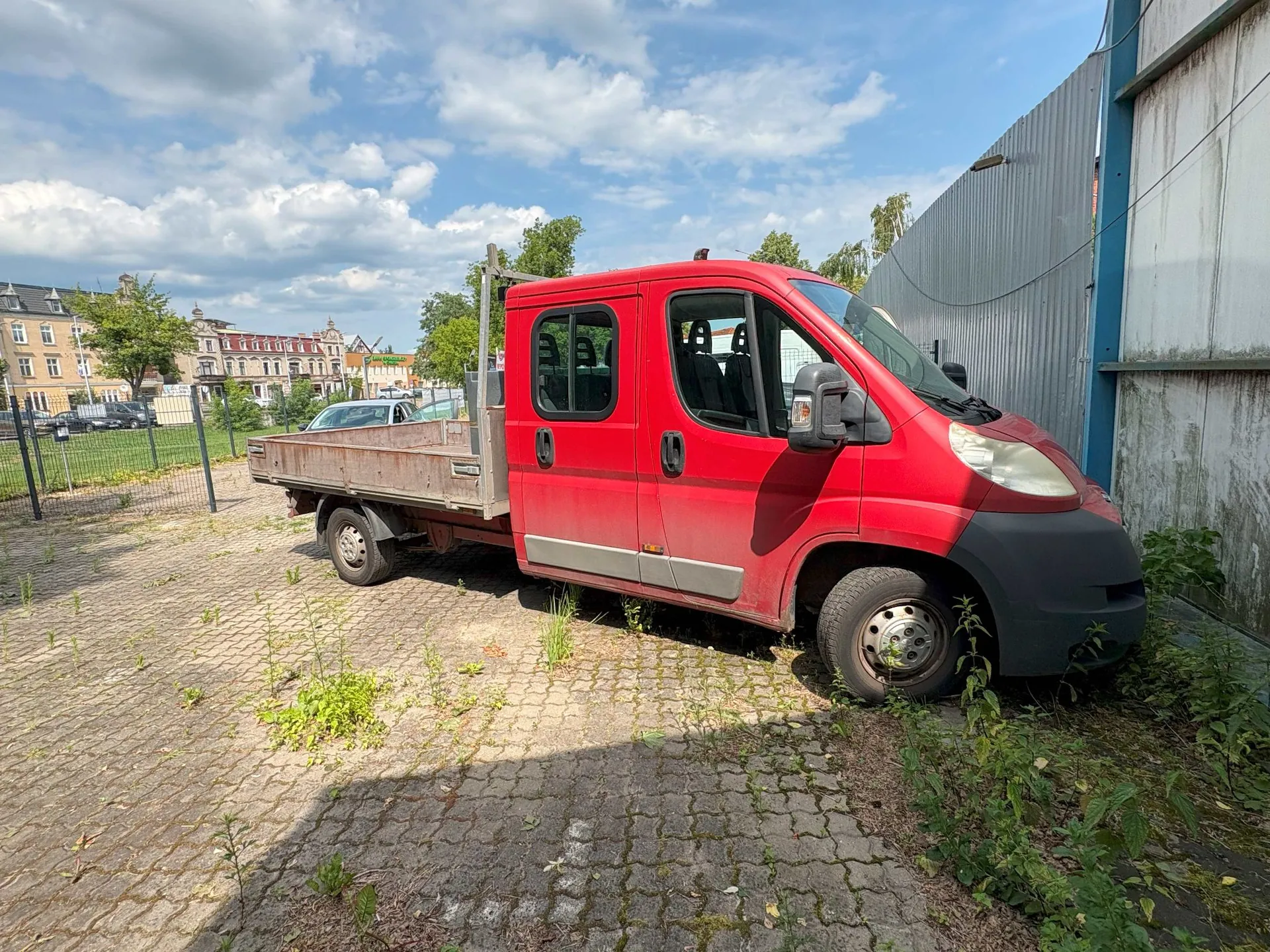 Czerwony samochód dostawczy typu platforma zaparkowany na brukowanym placu obok szarego budynku przemysłowego. Pojazd ma podwójną kabinę i otwartą przestrzeń ładunkową z tyłu. W tle widoczne są budynki mieszkalne, drzewa oraz niebo z białymi chmurami.