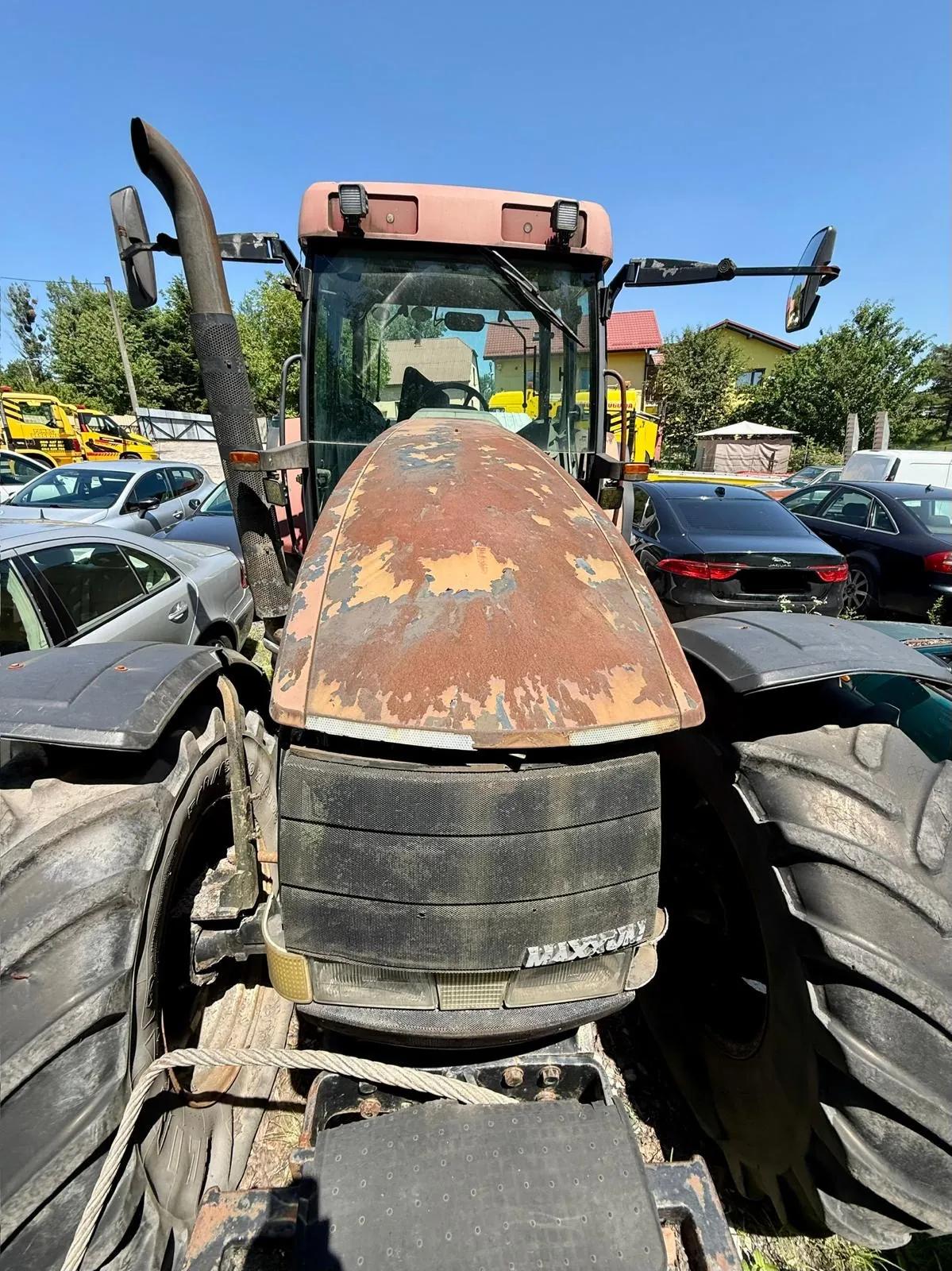 Zbliżenie na przód starego, czerwonego traktora z mocno zardzewiałą maską. Traktor posiada duże, czarne opony z wyraźnym bieżnikiem. Na przednim grillu widoczny jest napis 'MASSEY'. W tle widać zaparkowane samochody osobowe, domy oraz bezchmurne, niebieskie niebo.