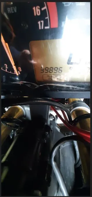 Zbliżenie na deskę rozdzielczą motocykla oraz fragmenty jego konstrukcji. W górnej części widoczny jest licznik z cyframi 16 i 17 oraz wyświetlacz LCD pokazujący przebieg 39896. Poniżej znajduje się widok na elementy mechaniczne, w tym złote amortyzatory i przewody.