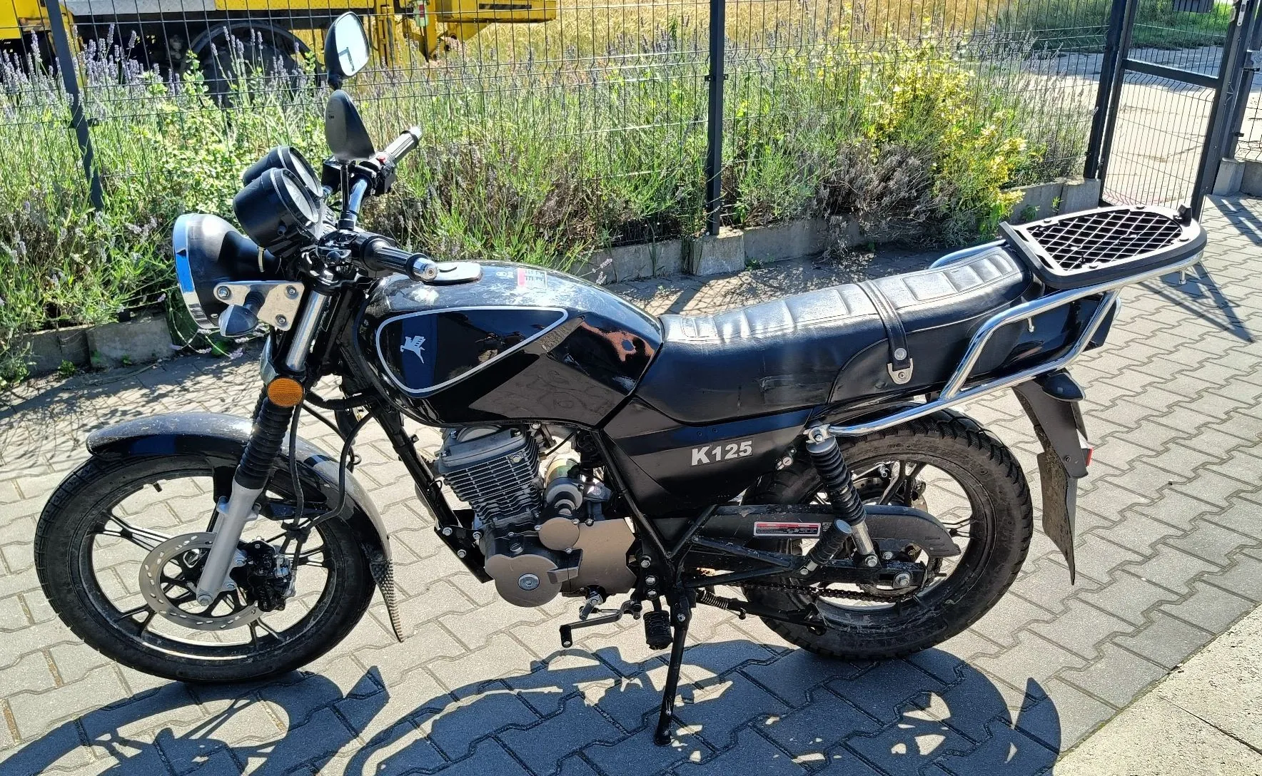 Czarny motocykl model K125 zaparkowany na kostce brukowej. Pojazd posiada okrągły reflektor, czarną kanapę z zamontowanym z tyłu bagażnikiem oraz srebrne elementy silnika i ramy. W tle widoczny jest metalowy płot, zieleń oraz fragment żółtego pojazdu.