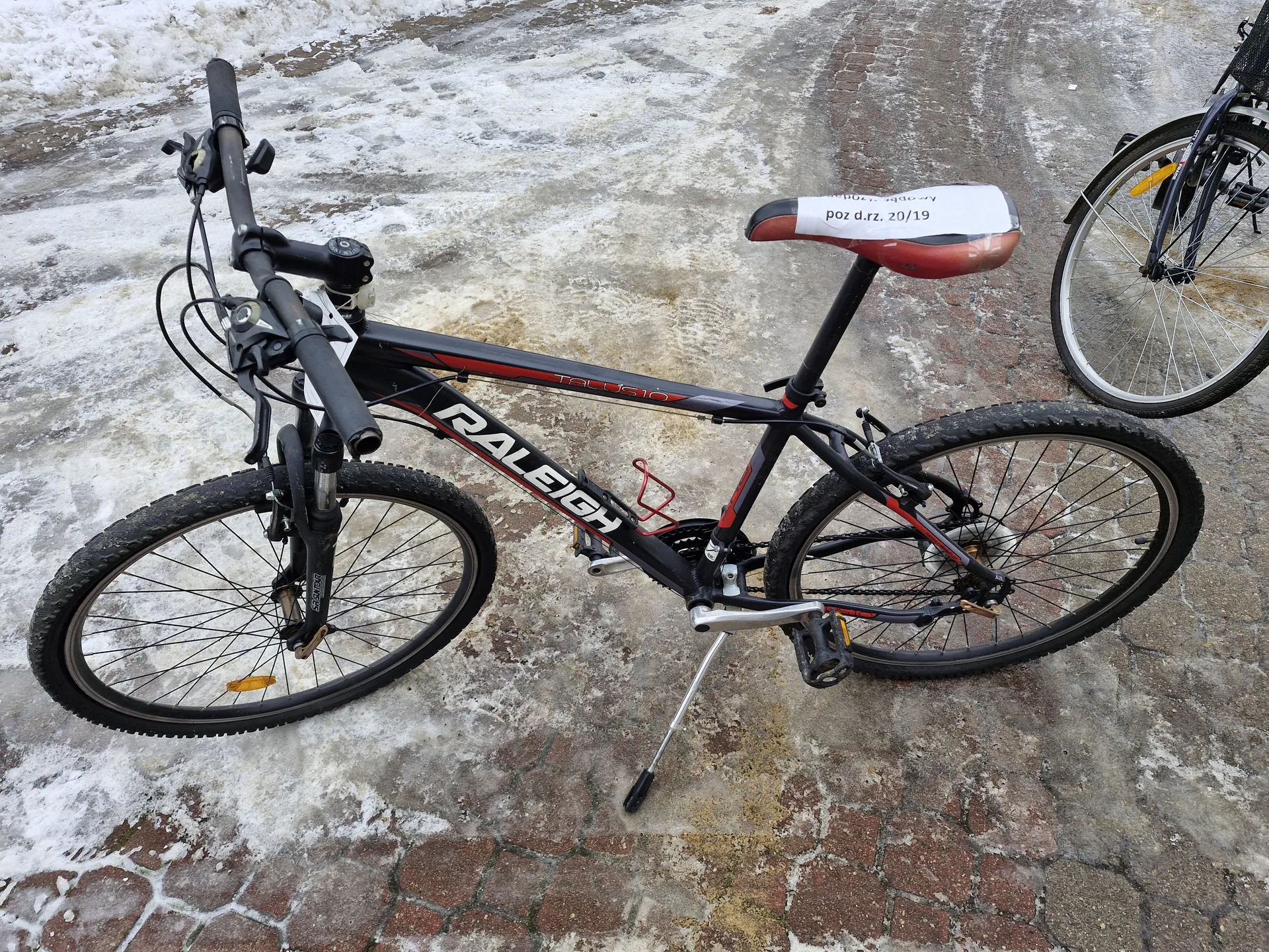 Czarny rower marki RALEIGH z czerwonymi akcentami, model TALUS 10, stojący na podkładowym stojaku na brukowanej nawierzchni z resztkami śniegu i lodu. Na siedzisku naklejony biały papier z napisem 'poz d.rz. 20/19'. Widoczne są czarne kierownica, koła z grubymi oponami oraz czerwony uchwyt na bidon. Po prawej stronie częściowo widoczny drugi rower z żółtym reflektorem na przednim kole.