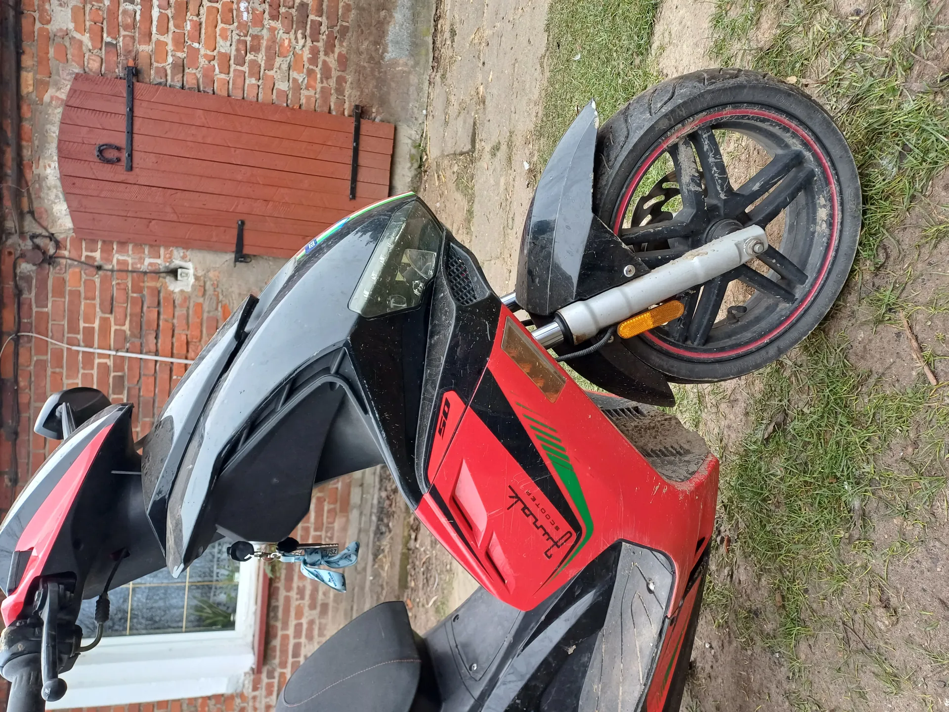 Czerwono-czarny skuter HONDA z napisem "50" i zielonymi akcentami, zaparkowany na brudnej ścieżce z trawą. Widoczne zniszczone opony z czerwonymi obręczami, niebieskie klucze, ceglana ściana z czerwonymi drzwiami i oknem. Brudna karoseria.