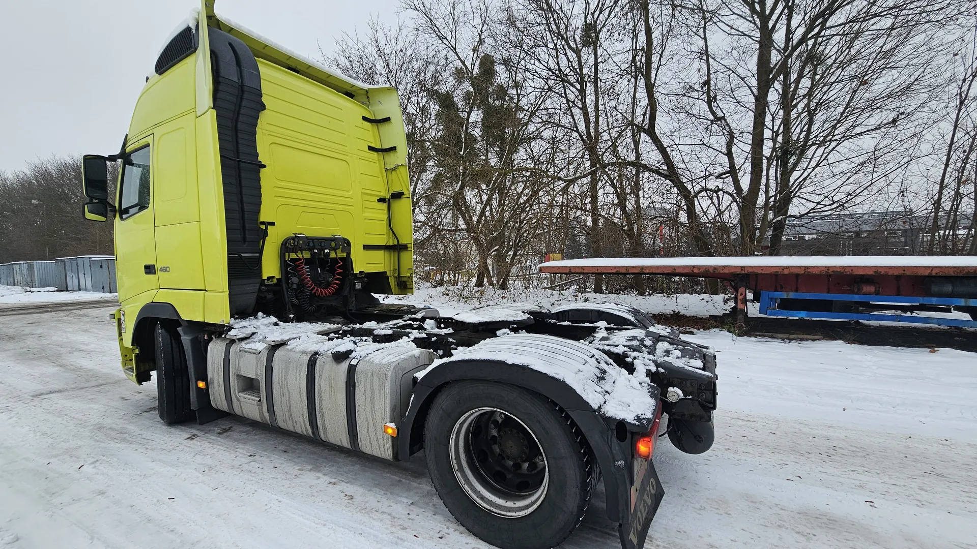 Żółta ciężarówka Volvo 460 z napisem "460" na boku, bez naczepy, pokryta śniegiem, stoi na śnieżnej drodze. W tle bezlistne drzewa, czerwono-niebieska naczepa z śniegiem, przemysłowe kontenery. Śnieg na zbiornikach paliwa, oponach i włączonych pomarańczowych światłach.