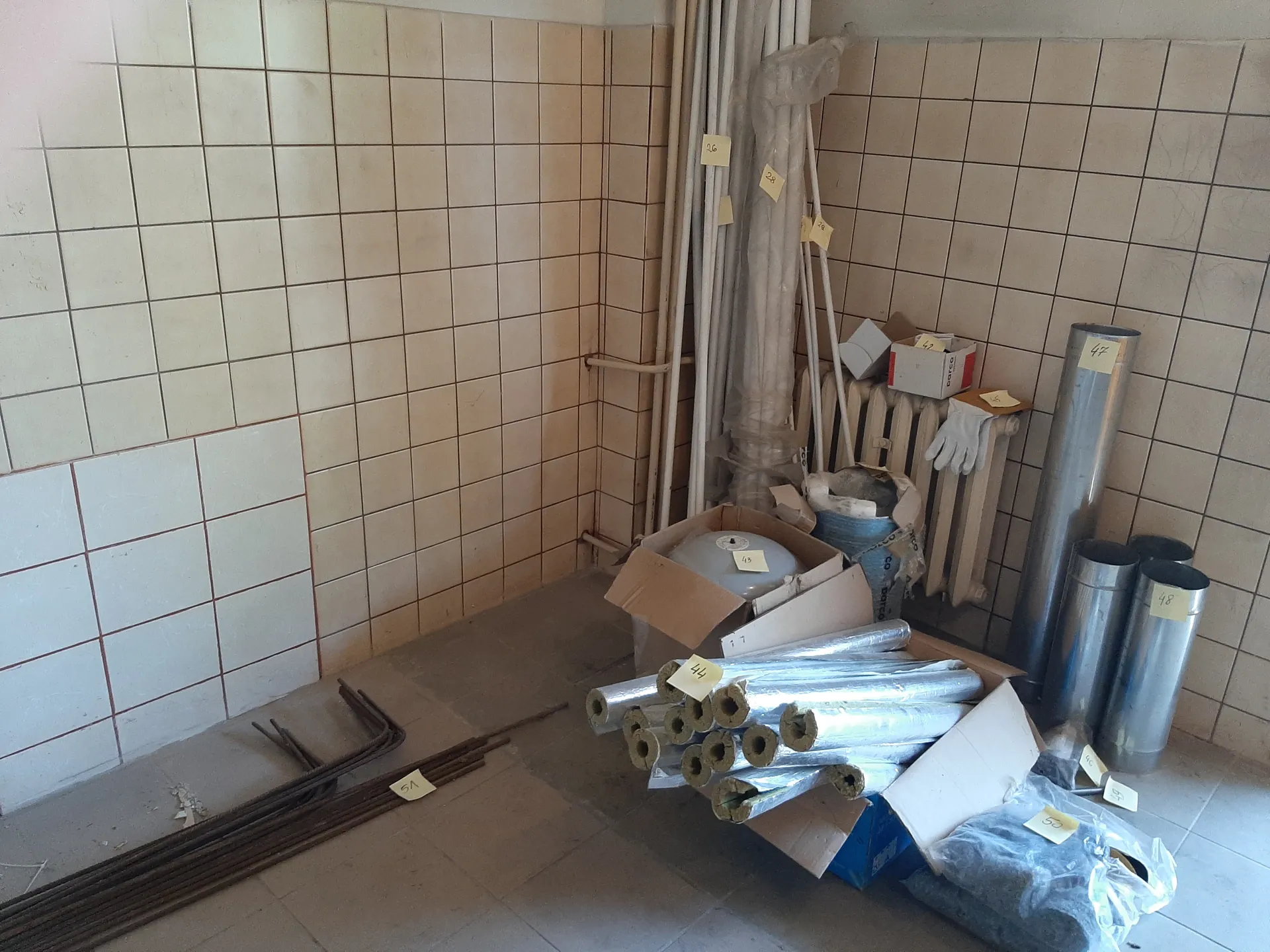 Pomieszczenie z beżowymi i białymi kafelkami na ścianach. Na podłodze leżą metalowe pręty z etykietą 5A, rolki izolacji lub rur owinięte folią z etykietą 44, karton z białym przedmiotem oznaczonym 43, metalowe rury z etykietami 48 i 49, biały grzejnik z rękawiczkami, pudełka oraz opakowania w folii. W tle widoczne są długie rury owinięte folią z żółtymi etykietami.