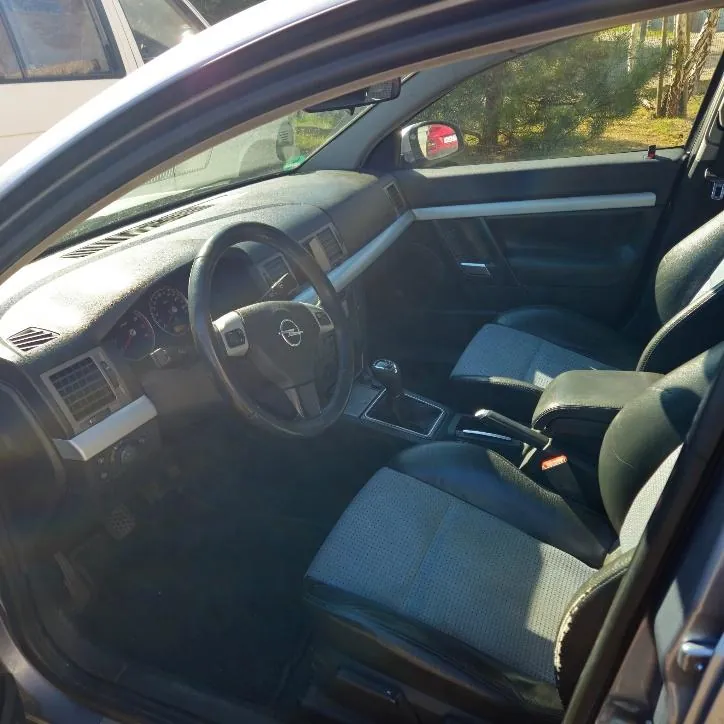 Wnętrze samochodu marki Nissan widziane z otwartego drzwi pasażera. Widoczna kierownica z logo Nissan, deska rozdzielcza z wskaźnikami, czarne siedzenia z szarym materiałem w środku, dźwignia zmiany biegów i hamulec ręczny. Przez otwarte drzwi widać fragment zewnętrzny z drzewami i innym pojazdem na tle. Światło słoneczne wpada do wnętrza, powodując lekkie odblaski na desce rozdzielczej.