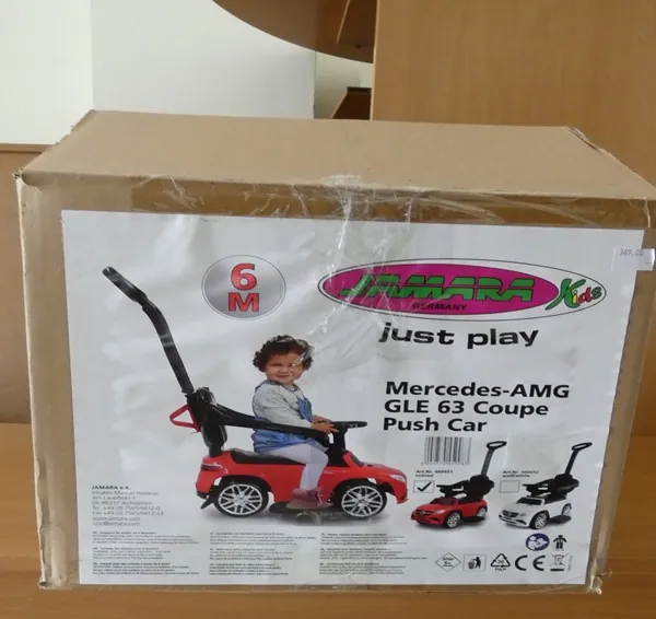 Kartonowa pudełko z opakowaniem produktu. Na etykiecie widoczny czerwony wózek dla dzieci w kształcie Mercedes-AMG GLE 63 Coupe Push Car z czarnym siedzeniem i uchwytem. Obok siedzi dziecko w niebieskiej koszuli i szarych spodniach. W górnej części etykiety znajduje się logo "Jamara Germany" z napisem "just play" oraz okrągły znak "6 M". Poniżej głównego obrazu umieszczono mniejsze zdjęcia wózka w kolorach czerwonym i białym. Na dole etykiety widoczne są symbole certyfikacyjne (w tym CE), kod kreskowy oraz dodatkowy tekst w języku niemieckim. Opakowanie jest owinięte przezroczystą folią.