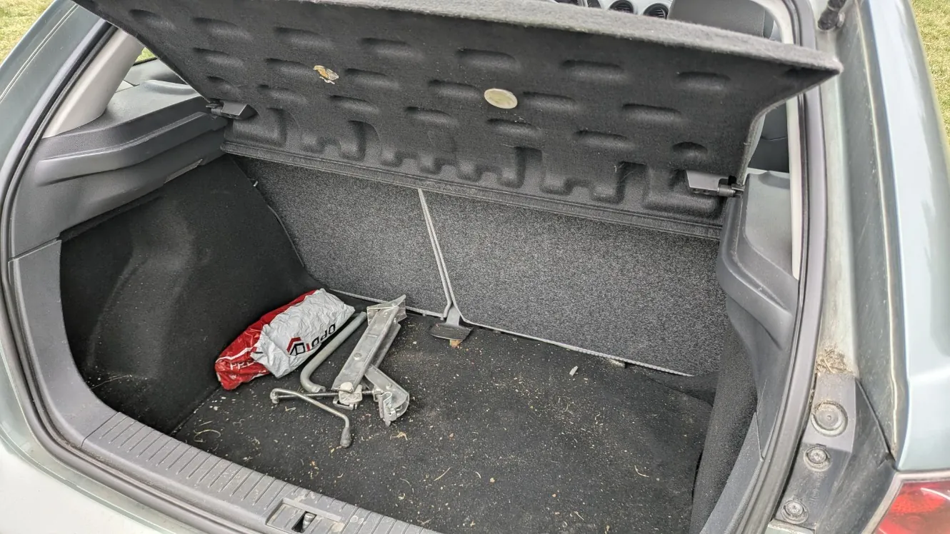 Otwarty bagażnik samochodu z czarnym wykładziny. Wewnątrz znajduje się metalowy podnośnik, czerwono-szara torba z widocznym tekstem 'DRO' oraz 'FRE' oraz kilka drobnych śmieci. Bagażnik posiada szarą obudowę, a w tle widoczna jest trawa. Samochód ma szarą karoserię.