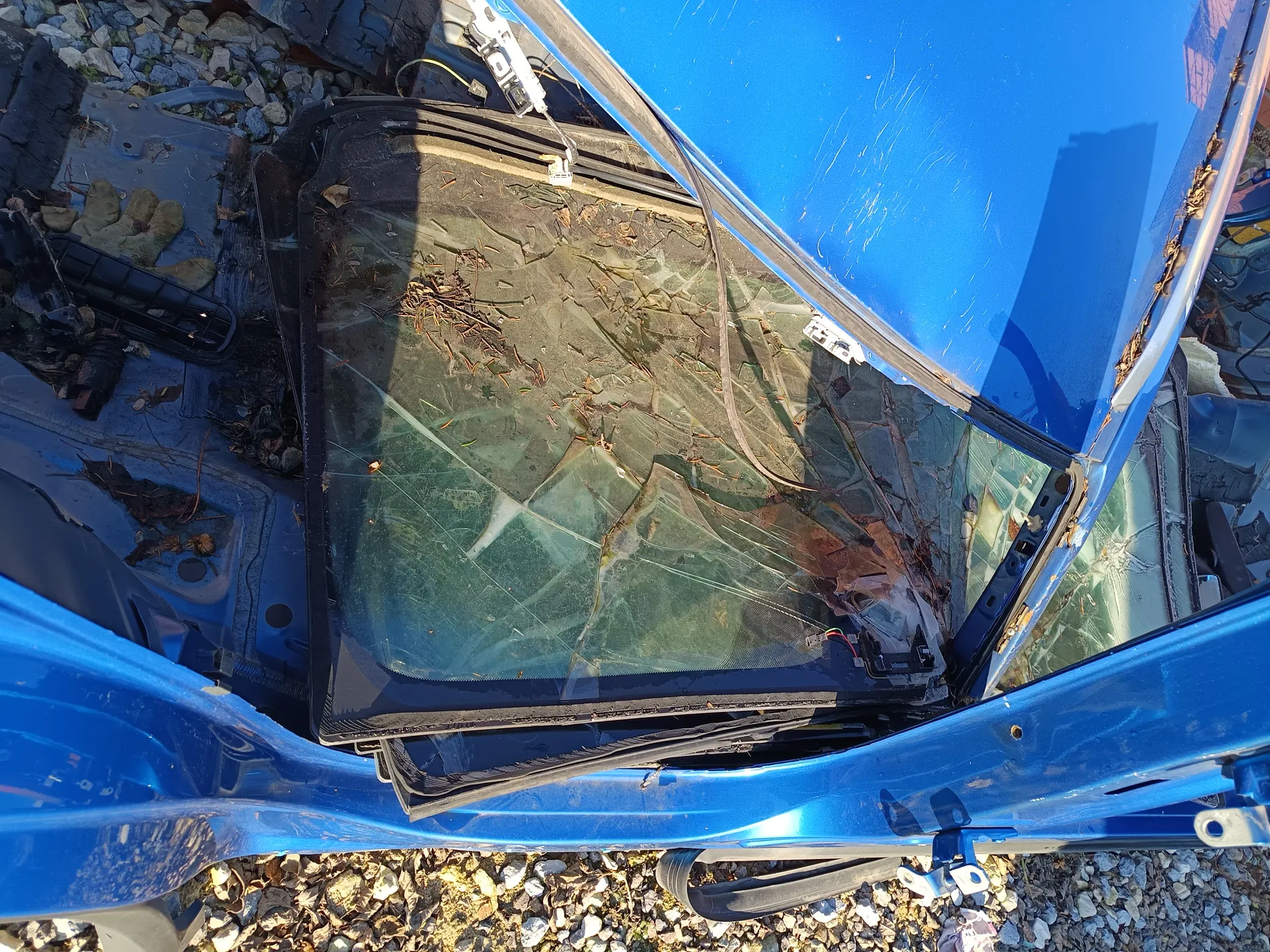 Zniszczony niebieski samochód z pękniętym przednim szybą. Szyba jest rozbita na wiele kawałków, na niej widoczne są liście i śmieci. Samochód znajduje się na żwirowym podłożu, wokół rozrzucone są części pojazdów i inne śmieci.