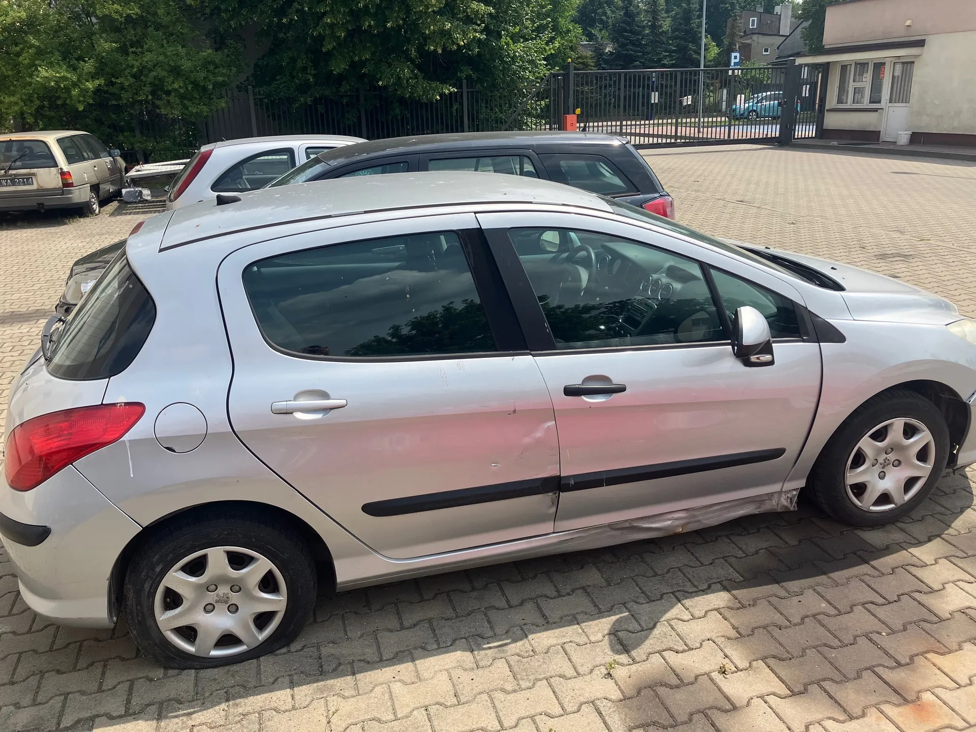 Srebrny samochód osobowy marki Opel Astra w nadwoziu hatchback, zaparkowany bokiem na kostce brukowej. Pojazd posiada widoczne uszkodzenia w postaci wgnieceń na dolnej części tylnych i przednich drzwi od strony pasażera. W tle widoczne są inne zaparkowane samochody, czarny metalowy płot oraz fragmenty budynków i zielone drzewa.