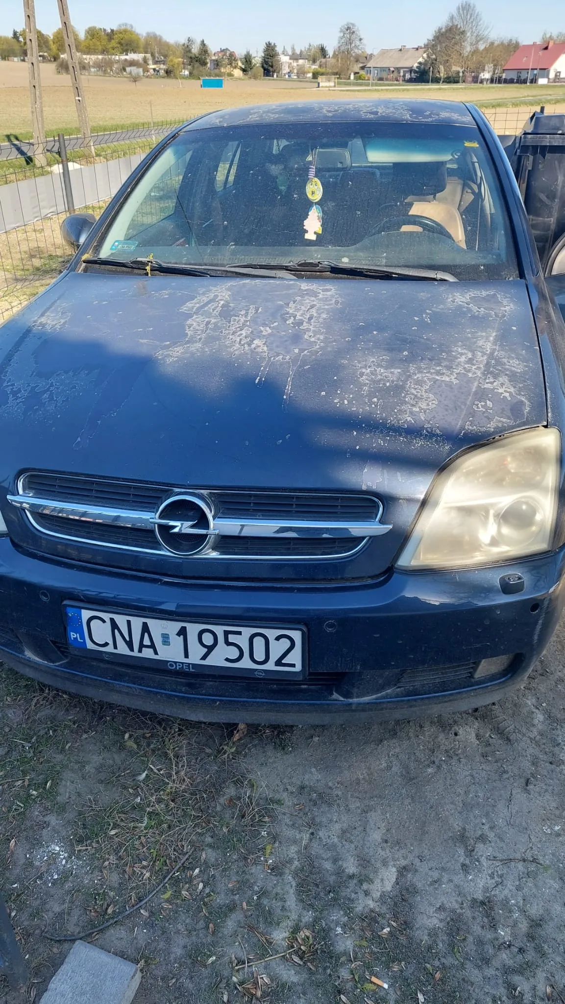 Przedni widok niebieskiego samochodu marki Opel z tablicą rejestracyjną CNA 19502 (PL). Na masce widoczne są białe plamy, prawdopodobnie od ptaków. Wewnątrz kabiny na szybie wiszą dekoracje: żółty emotikon i mała figurka. Samochód stoi na podłożu z trawą i ziemią, w tle ogrodzenie, pola, domy i drzewa. Na tablicy rejestracyjnej widoczny napis "OPEL" pod numerem.