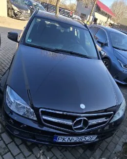 Czarny samochód marki Mercedes-Benz z widocznym logo na atrapie chłodnicy. Na przednim zderzaku umieszczona tablica rejestracyjna o numerze PK-N 2852F. Samochód zaparkowany na brukowanej powierzchni, w tle widoczne są inne pojazdy oraz budynki z czerwonym dachem i ogrodzeniem.