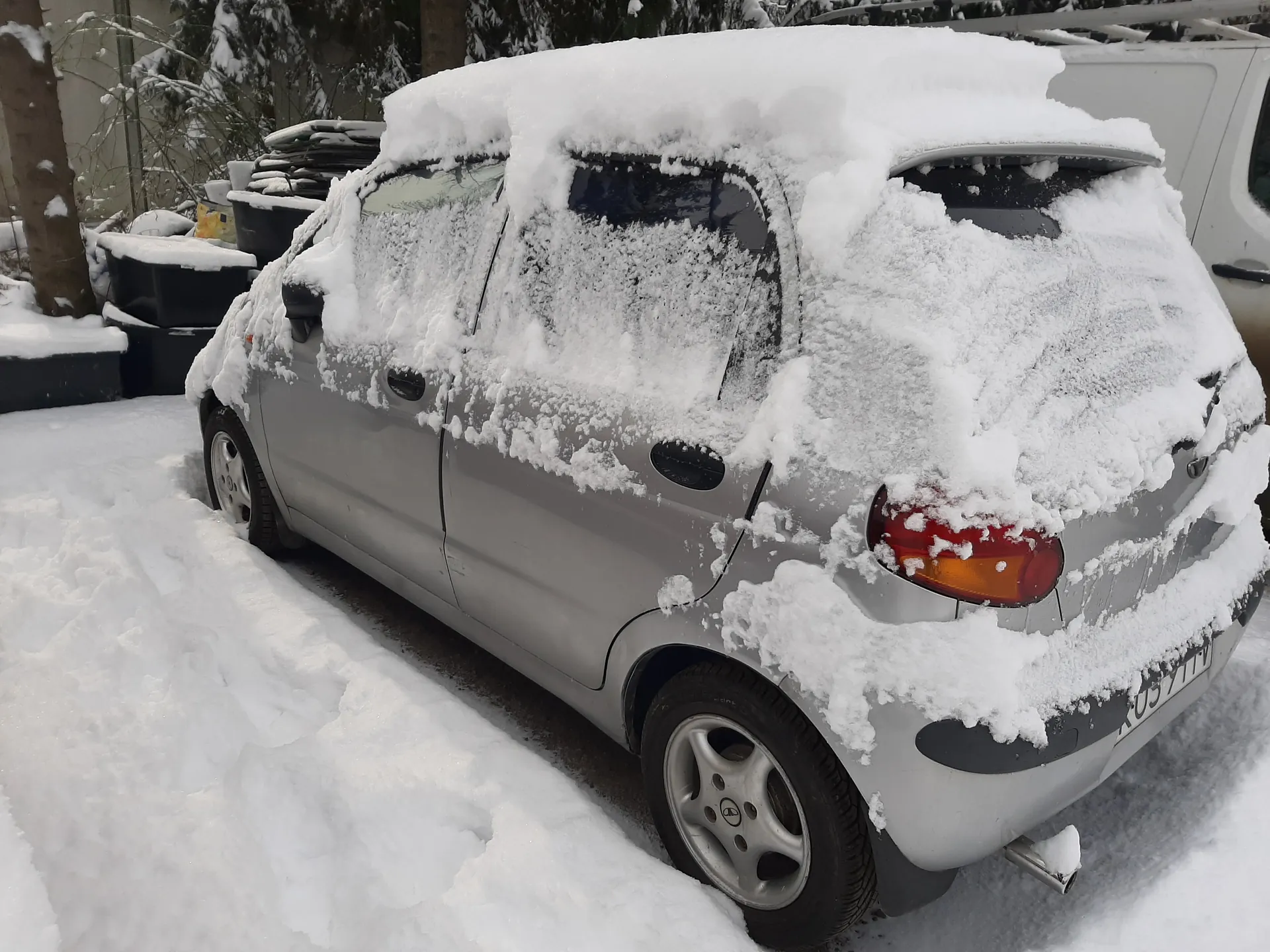 Mały srebrny samochód osobowy pokryty grubą warstwą śniegu, zaparkowany na śnieżnej powierzchni. Widoczne są zakryte śniegiem koła, tylne światła oraz fragment tablicy rejestracyjnej. W tle czarne pojemniki, drzewa i fragment białego pojazdu. Śnieg pokrywa również otaczający teren i elementy w tle.