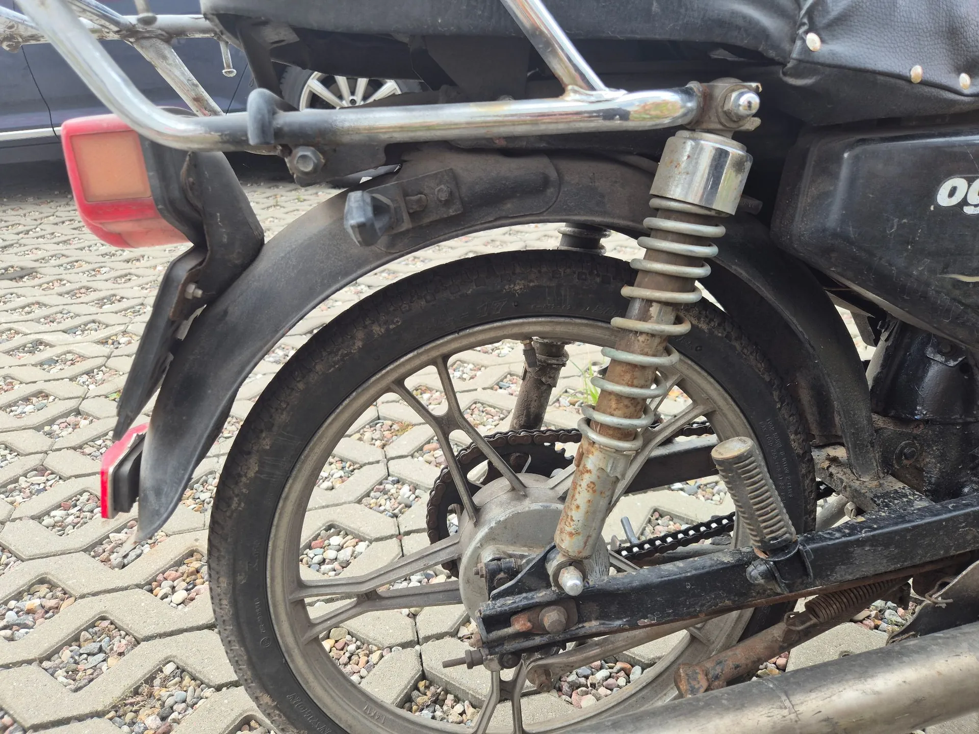 Zbliżenie na tylne koło i zawieszenie motocykla. Widoczna jest srebrna felga szprychowa, opona, łańcuch napędowy oraz amortyzator z widoczną sprężyną. Nad kołem znajduje się czarny błotnik i czerwone światło stopu. Motocykl stoi na kostce brukowej.